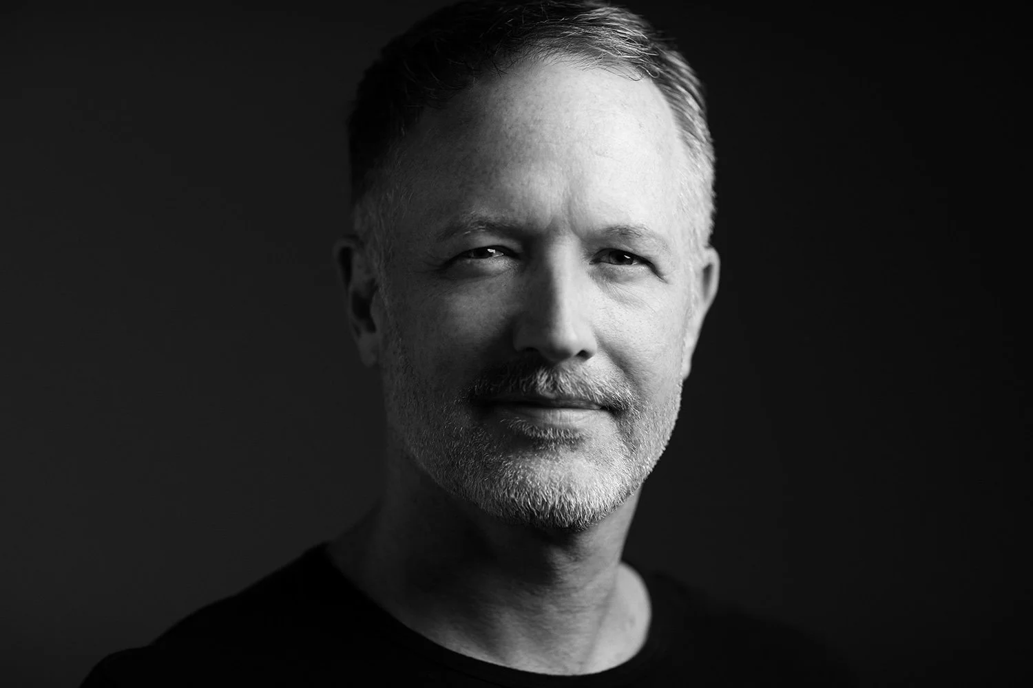 Black and white portrait of a middle-aged man with short, gray hair and a beard, smiling gently, against a dark background.