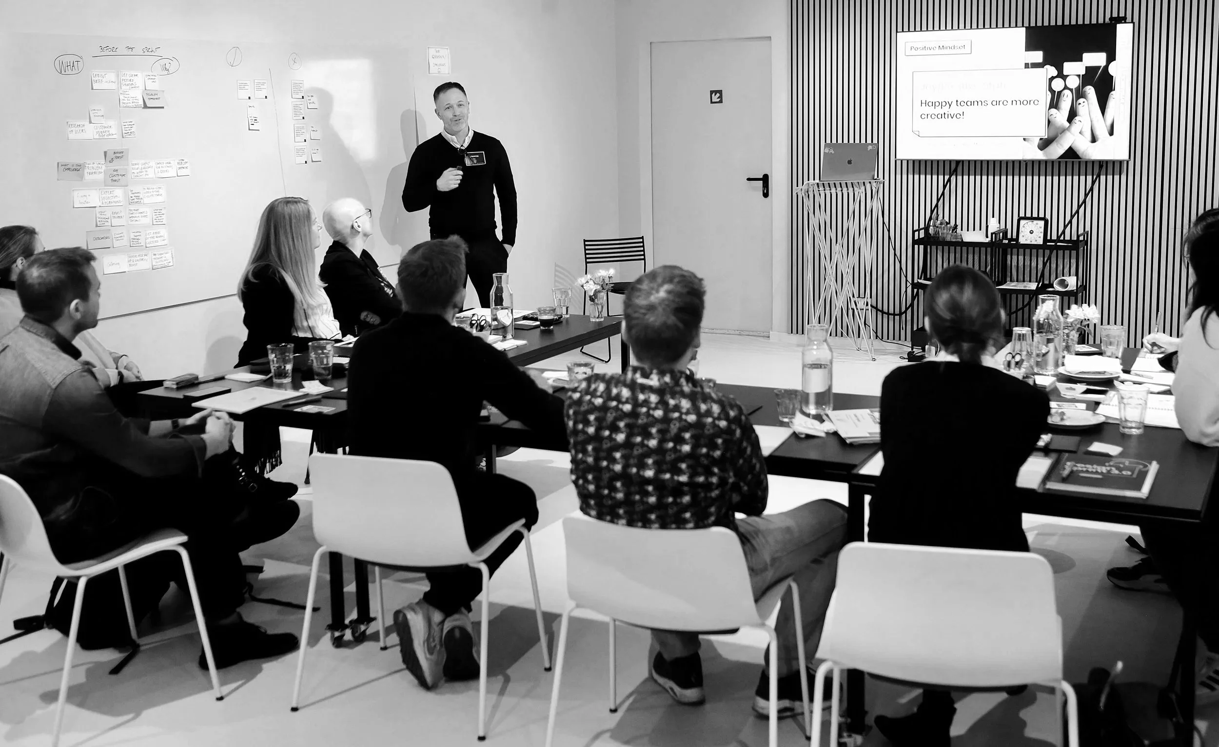 A black and white photo of a group of people attending a presentation in a modern conference room. The presenter, a man, is standing near a whiteboard with notes and diagrams, holding a microphone. The attendees are seated around a table with notebooks and glasses of water, listening to the presentation. A large screen displays a slide titled 'Positive Mindset' with the phrase 'Happy teams are more creative!'.