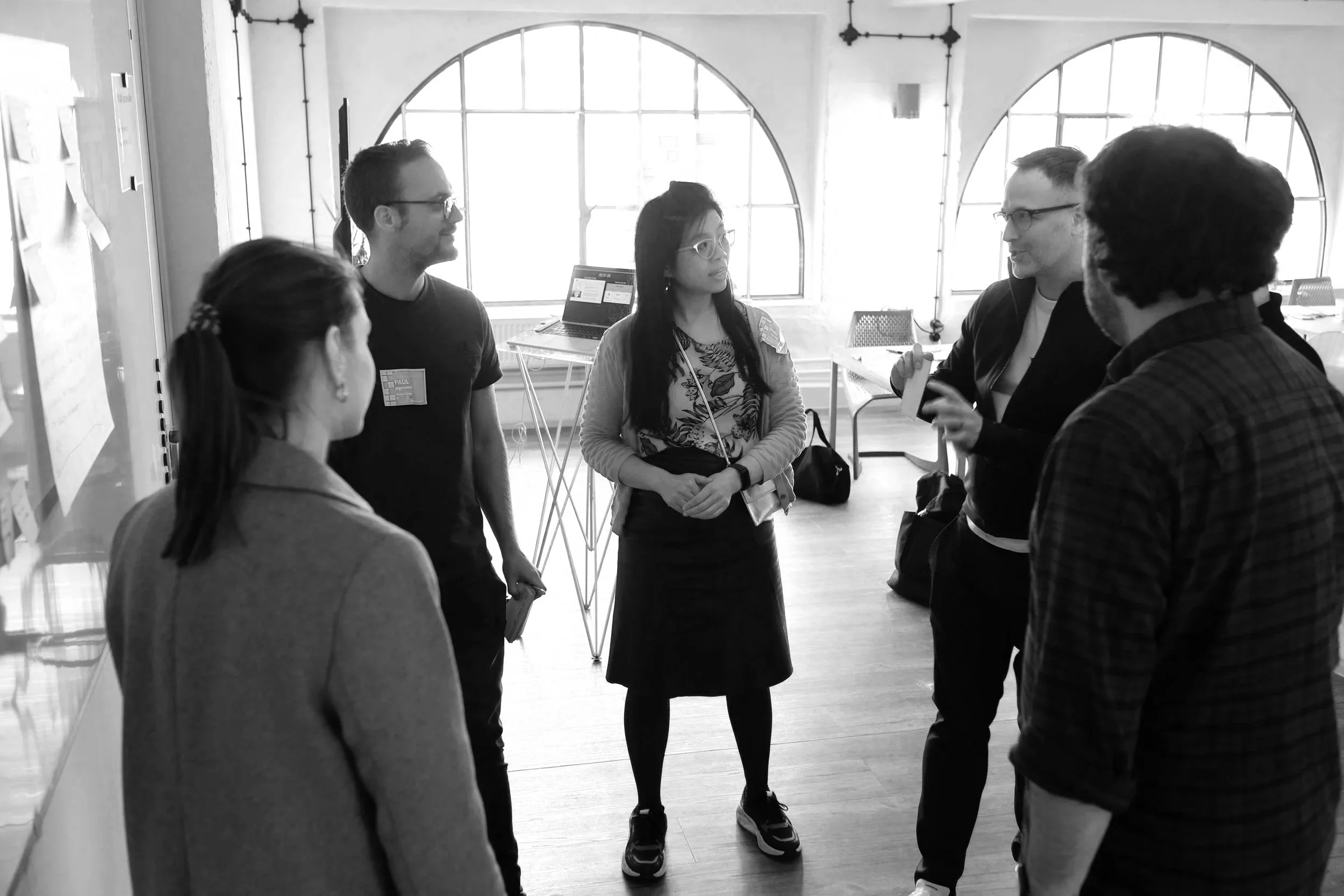 Five people in a circle having a discussion in a bright room with large windows and exposed brick walls.