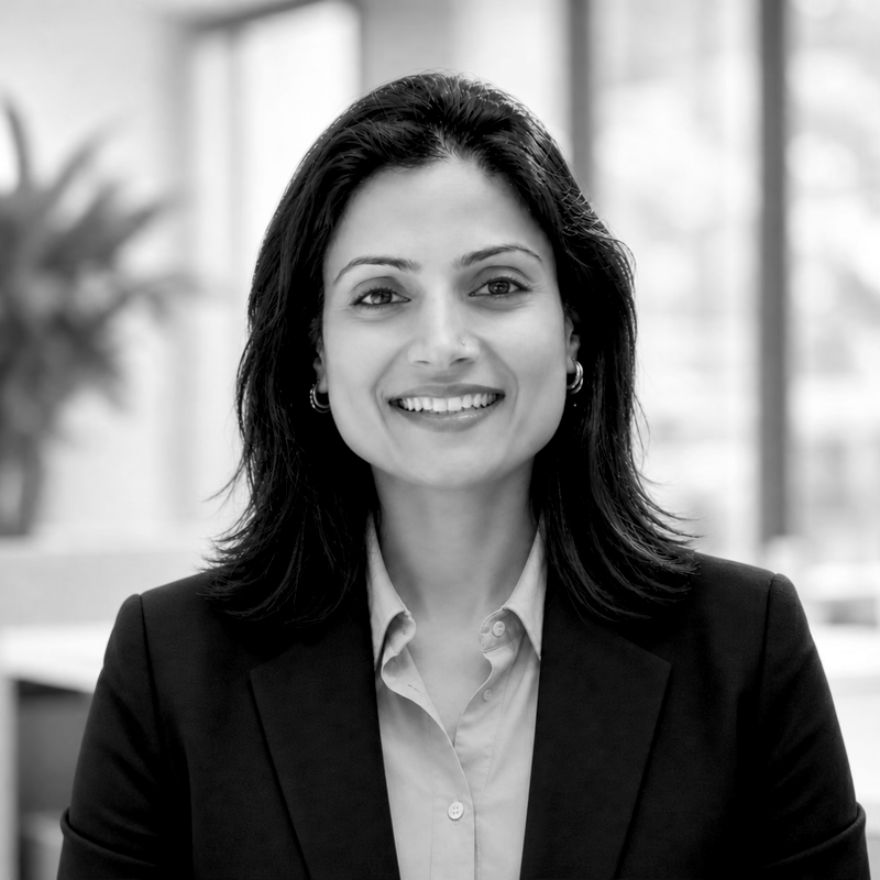 A woman with dark hair, wearing a blazer and a collared shirt, smiling in a professional setting with blurred background.