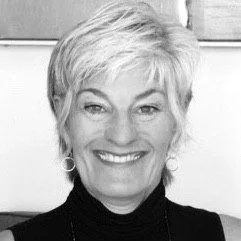 A portrait of a smiling woman with short white hair, wearing a dark sleeveless top and hoop earrings, in a black and white setting.