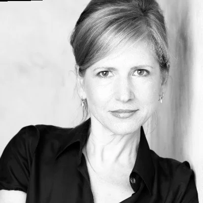 Black and white photo of a woman with a confident expression, wearing a dark shirt. Her hair is pulled back, and she is looking directly at the camera.