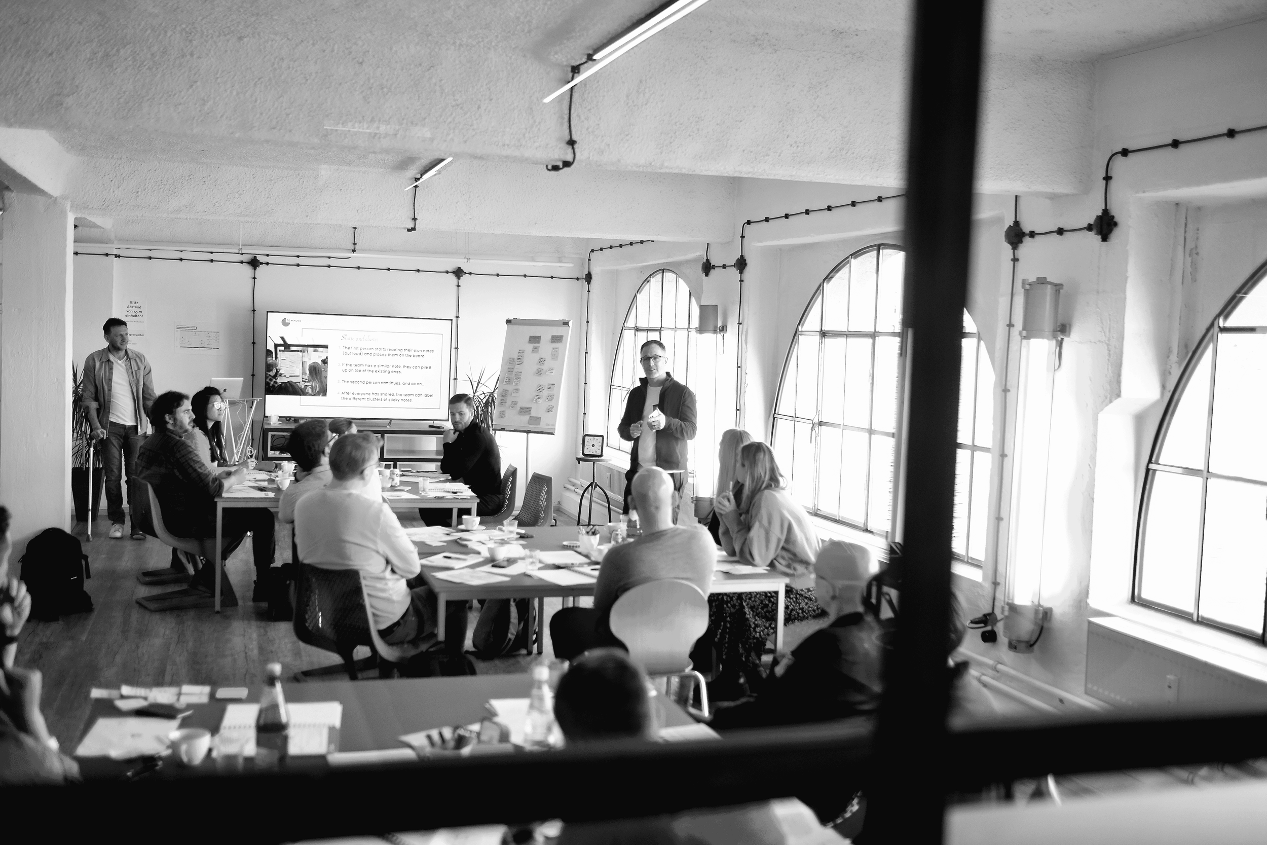 A black and white photo of a group of people attending a meeting or workshop in a bright room with large arched windows. One person is standing and speaking, while others are seated around tables, listening and taking notes. A large screen or monitor displays presentation slides.