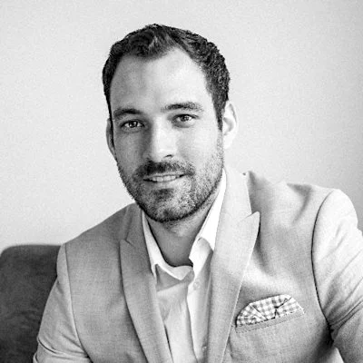 Black and white portrait of a man in a light-colored suit and open-collared shirt, sitting with a neutral expression.