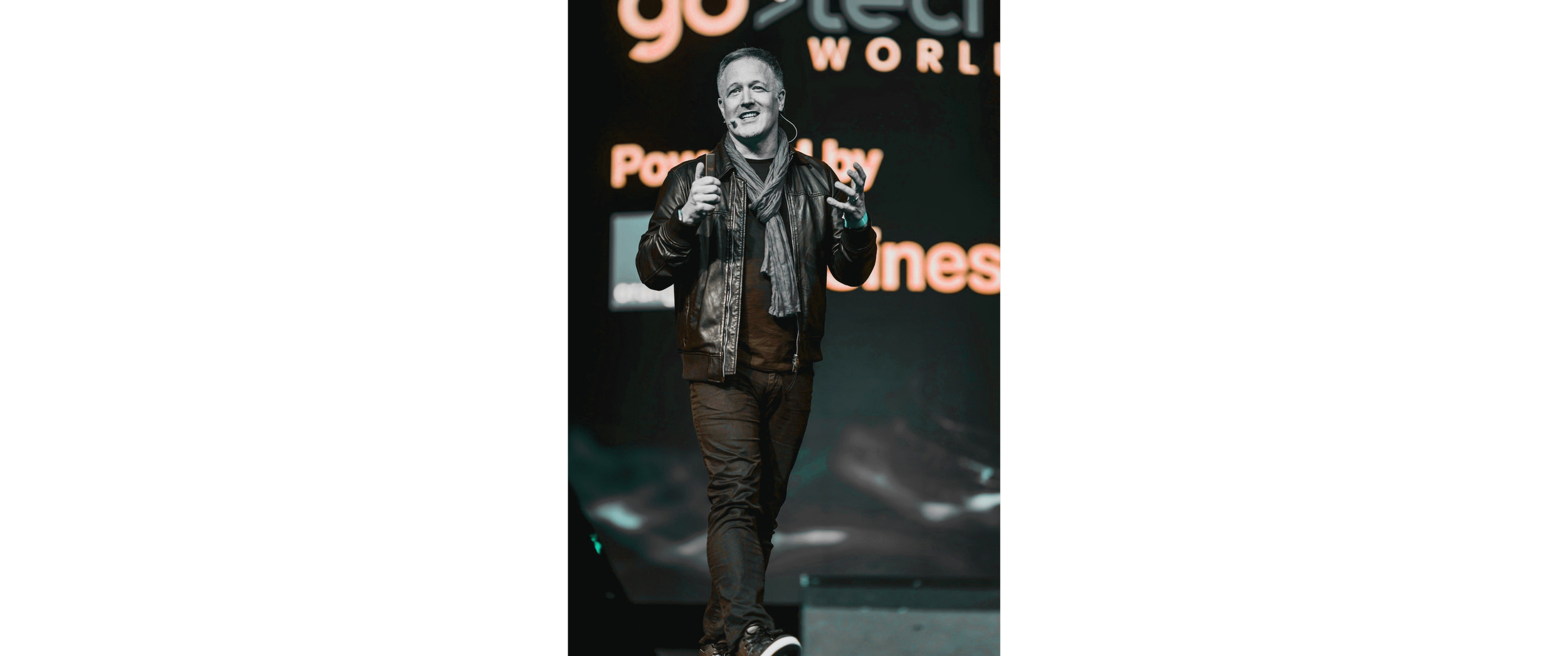 A man wearing a black leather jacket and scarf is speaking on stage with a microphone, with a background that has text including words like "goal" and "world".