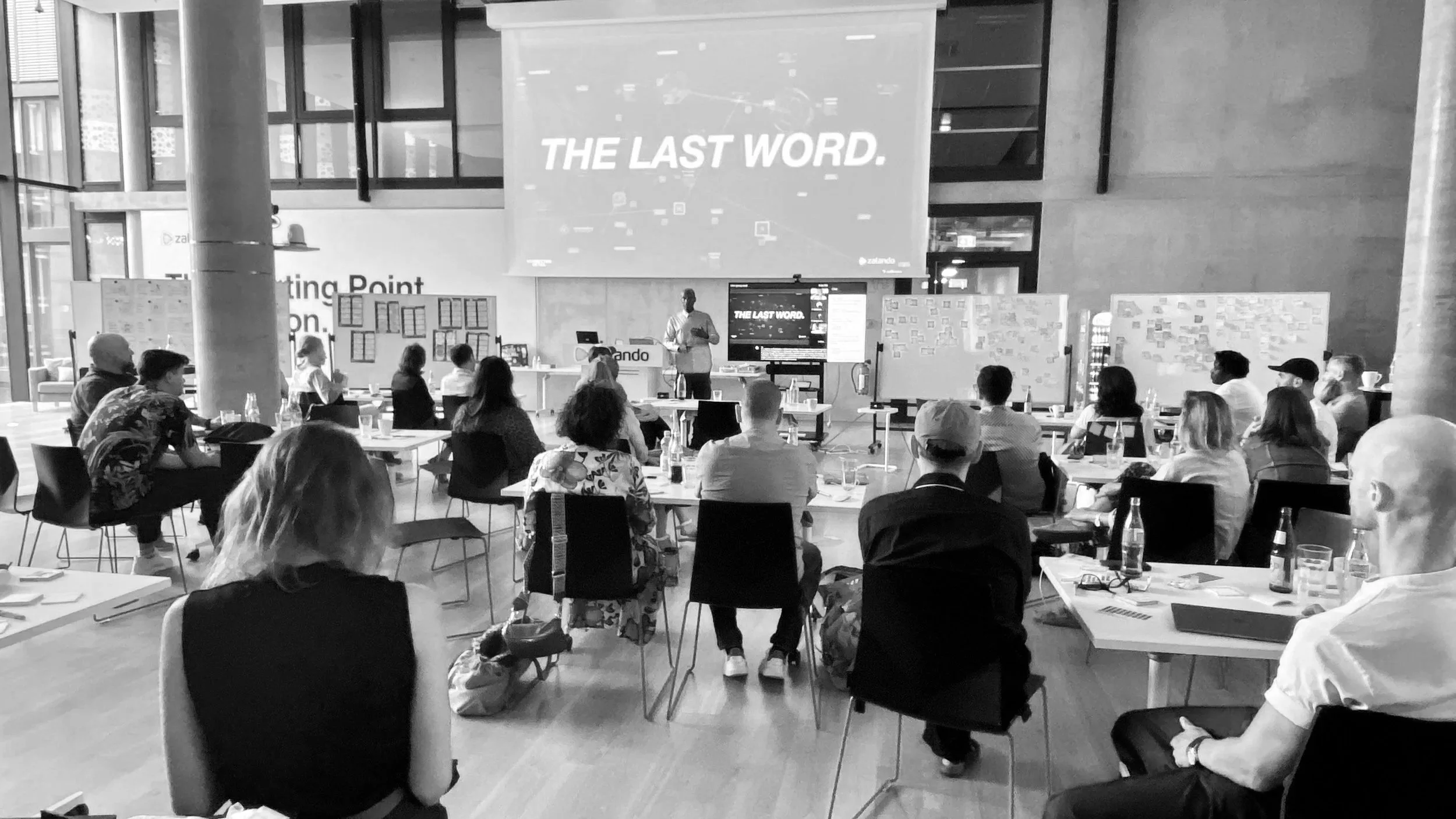A group of people seated at tables attending a presentation in a bright modern conference room, with a large screen displaying the words "The Last Word."