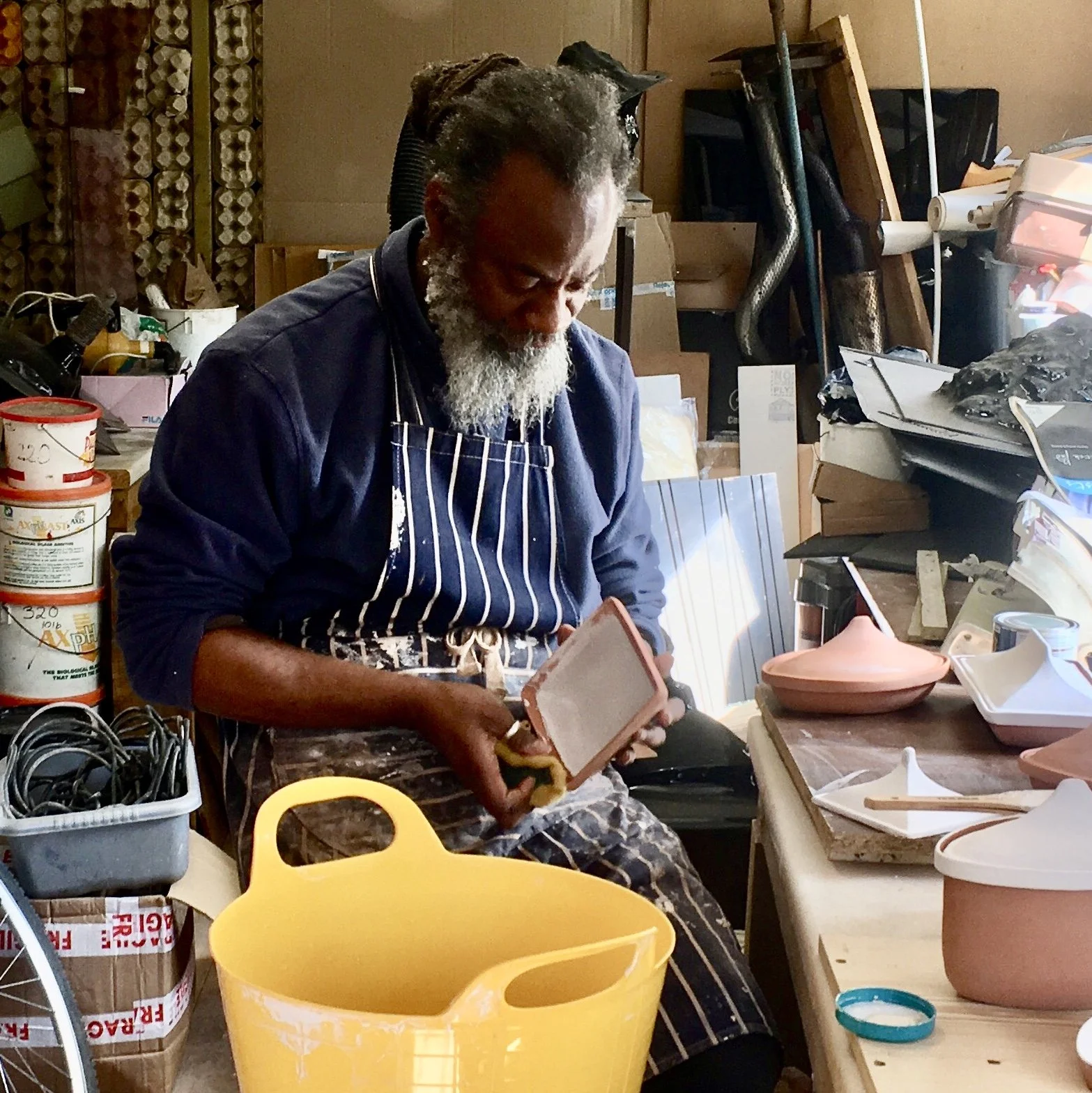 An older man with a gray beard, wearing a navy blue shirt and striped apron, working on a ceramic piece in a cluttered workshop surrounded by pottery and materials.