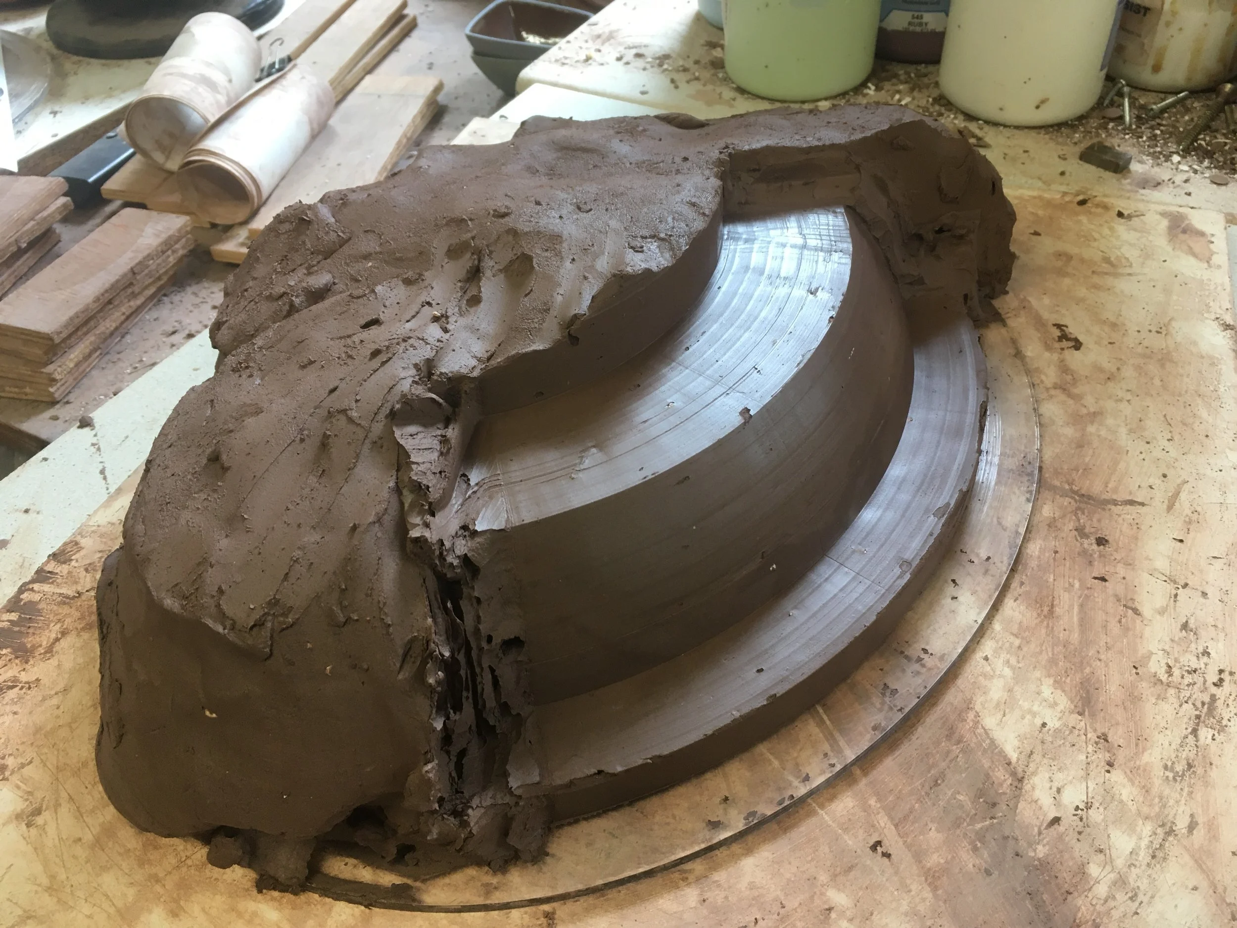 Clay mold in progress for pottery wheel, surrounded by pottery tools and supplies on a work table.