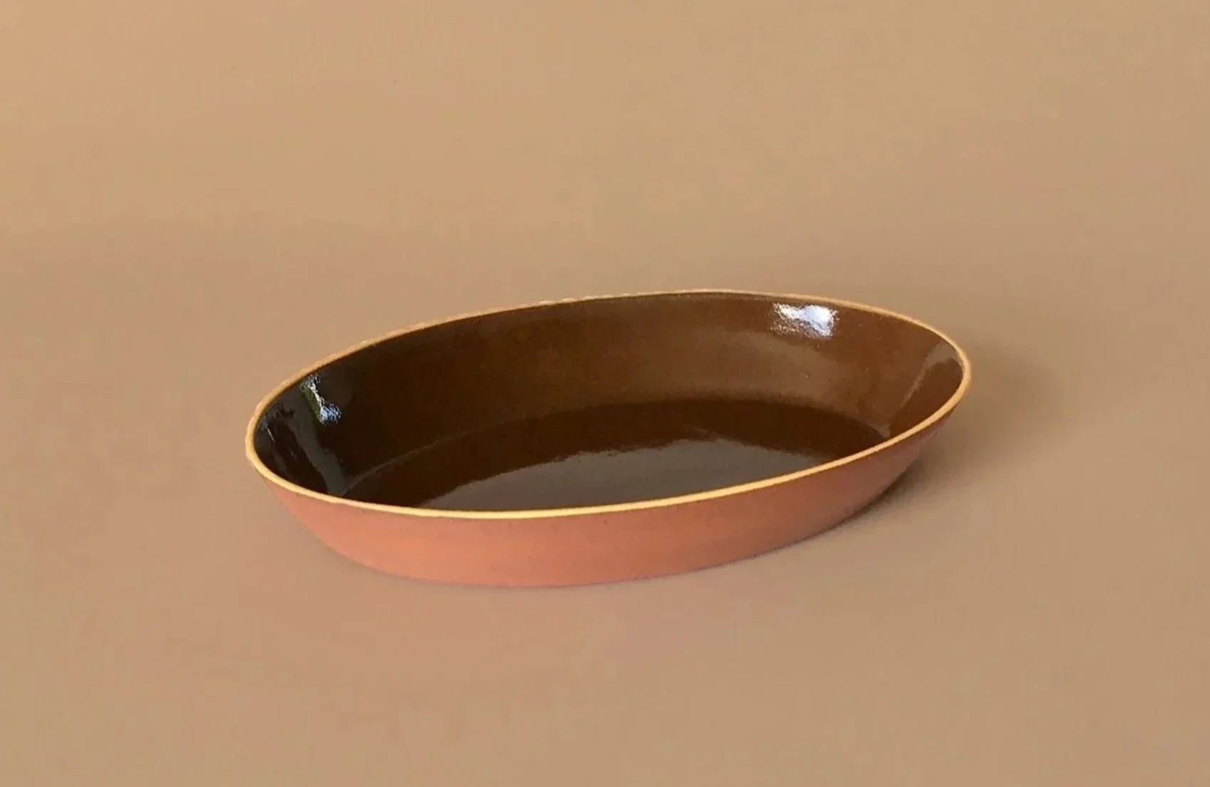 An oval-shaped ceramic dish with a glossy dark interior and a matte reddish exterior, placed on a beige surface.