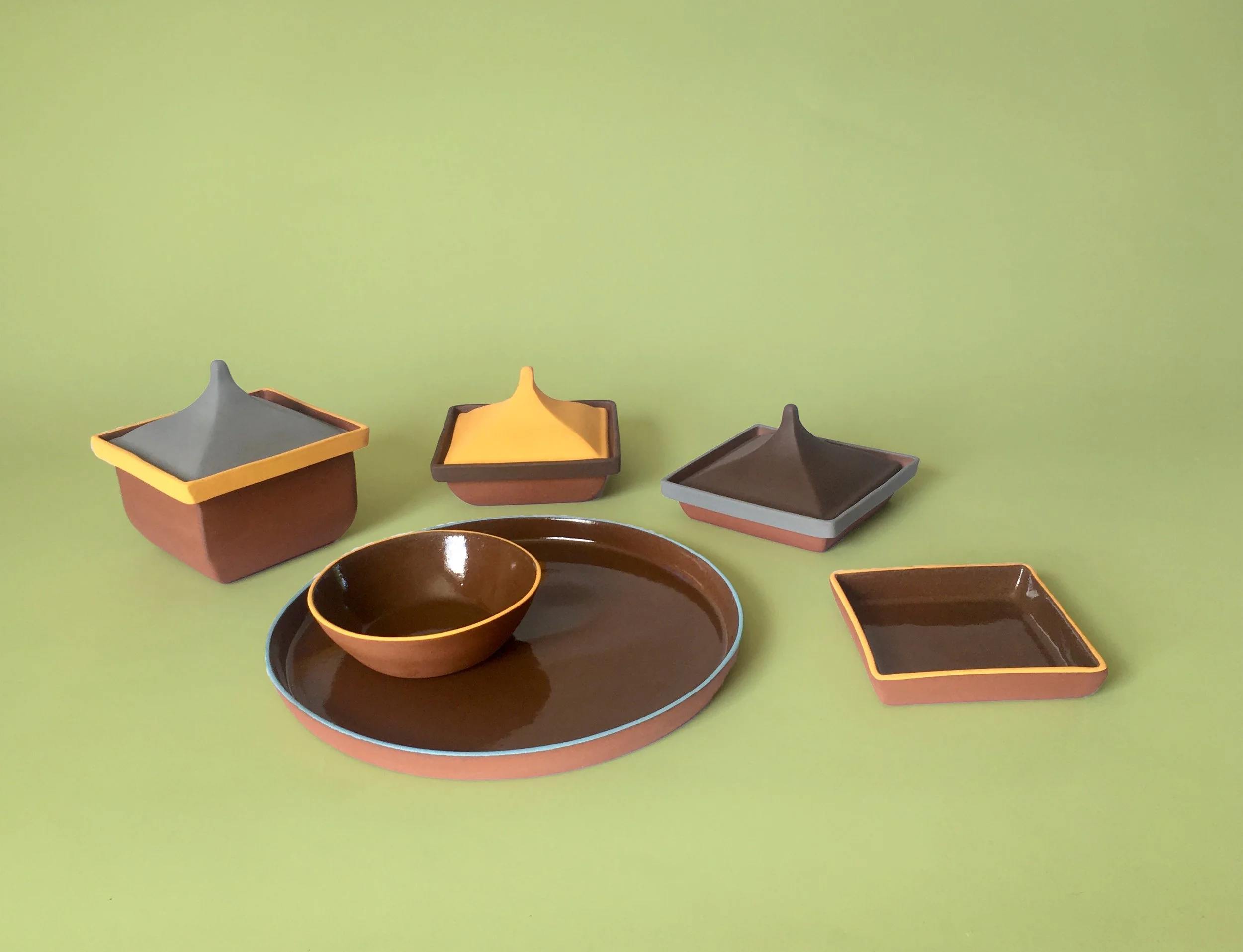 Set of six ceramic dishes with brown exteriors and colorful interiors against a green background, including a round plate with a smaller bowl, three square dishes, and two rectangular dishes, one with a lid.