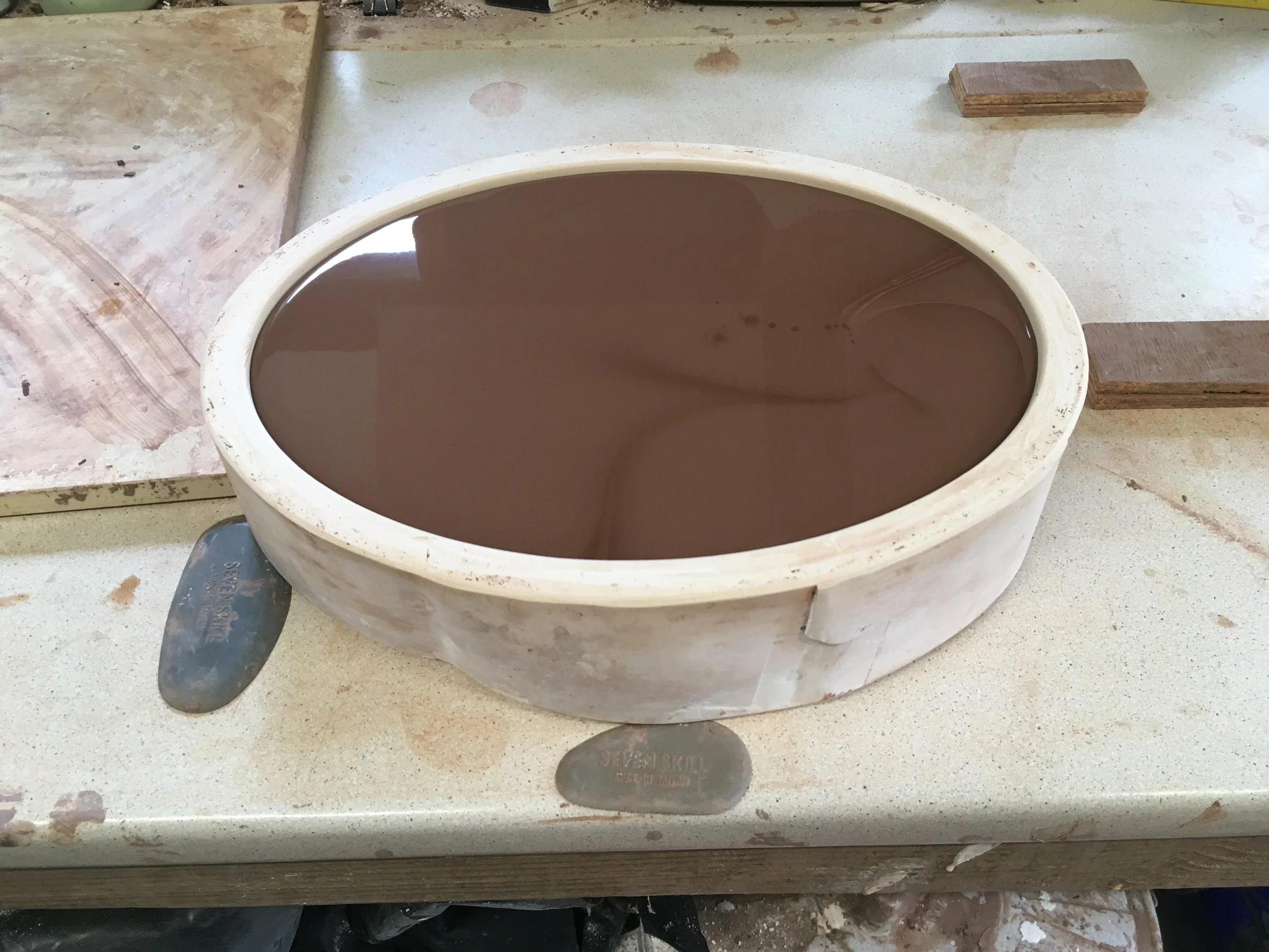 Ceramic basin filled with dark brown liquid, on a workshop table surrounded by wood pieces and tools.