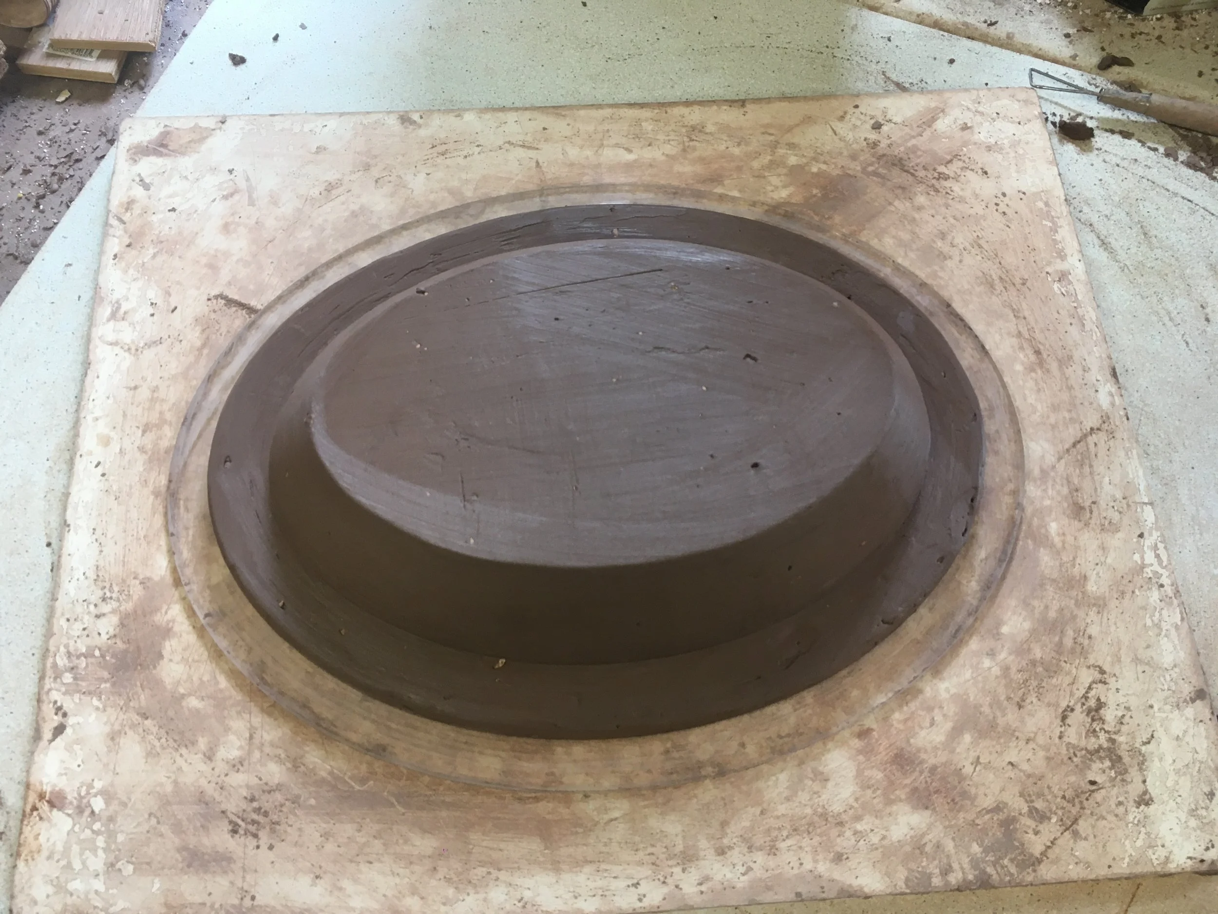 A rectangular piece of clay on a worktable with a circular depression in the center, forming a shallow bowl or dish shape, likely in the process of being shaped for pottery or ceramic work.