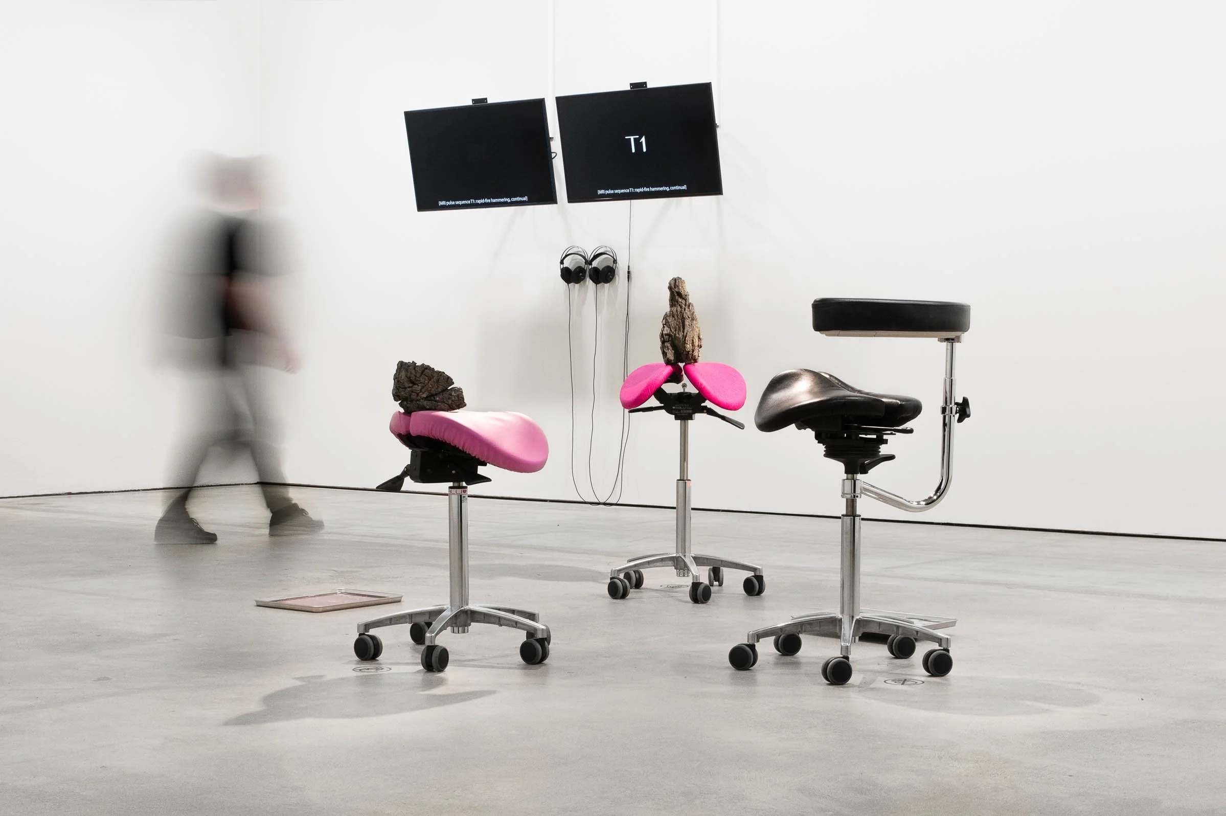 Foreground: Conflab (2023). 3 chunks of rock sit on split-seat saddle stools, ergonomic seats used by healthcare workers. The saddles are upholstered in pale pink, lurid pink and black. Image: Helen Korpak