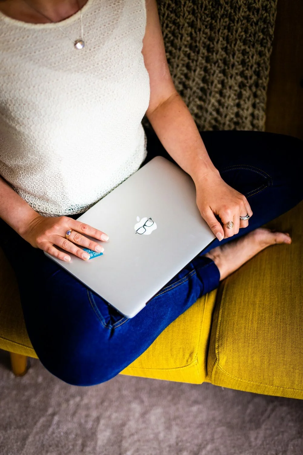A woman sitting on a yellow chair with her legs crossed, wearing a white textured top, blue jeans, and a ring on her left hand, holding a closed silver MacBook with a sticker of glasses on the lid.