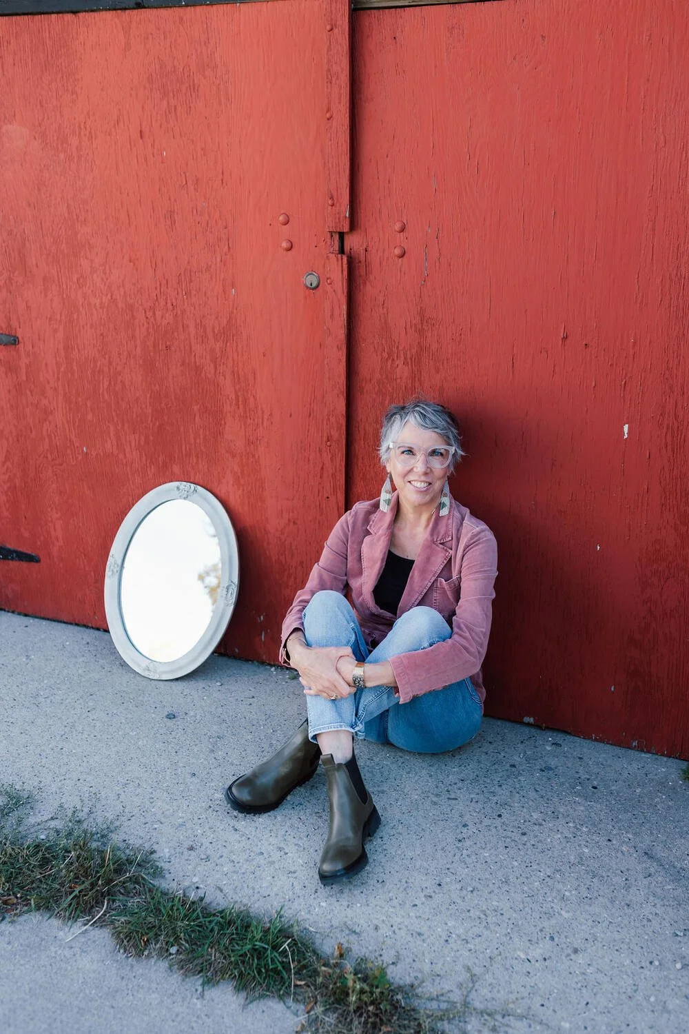 A woman with short gray hair and glasses sitting on the ground in front of a large red wooden wall, smiling at the camera, with a round mirror leaning against the wall beside her, and patches of grass and dirt on the sidewalk.