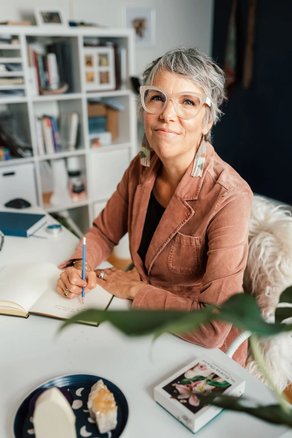 An older woman with short gray hair and glasses is sitting at a desk with a pen and open notebook, smiling at the camera in a home office or study space.