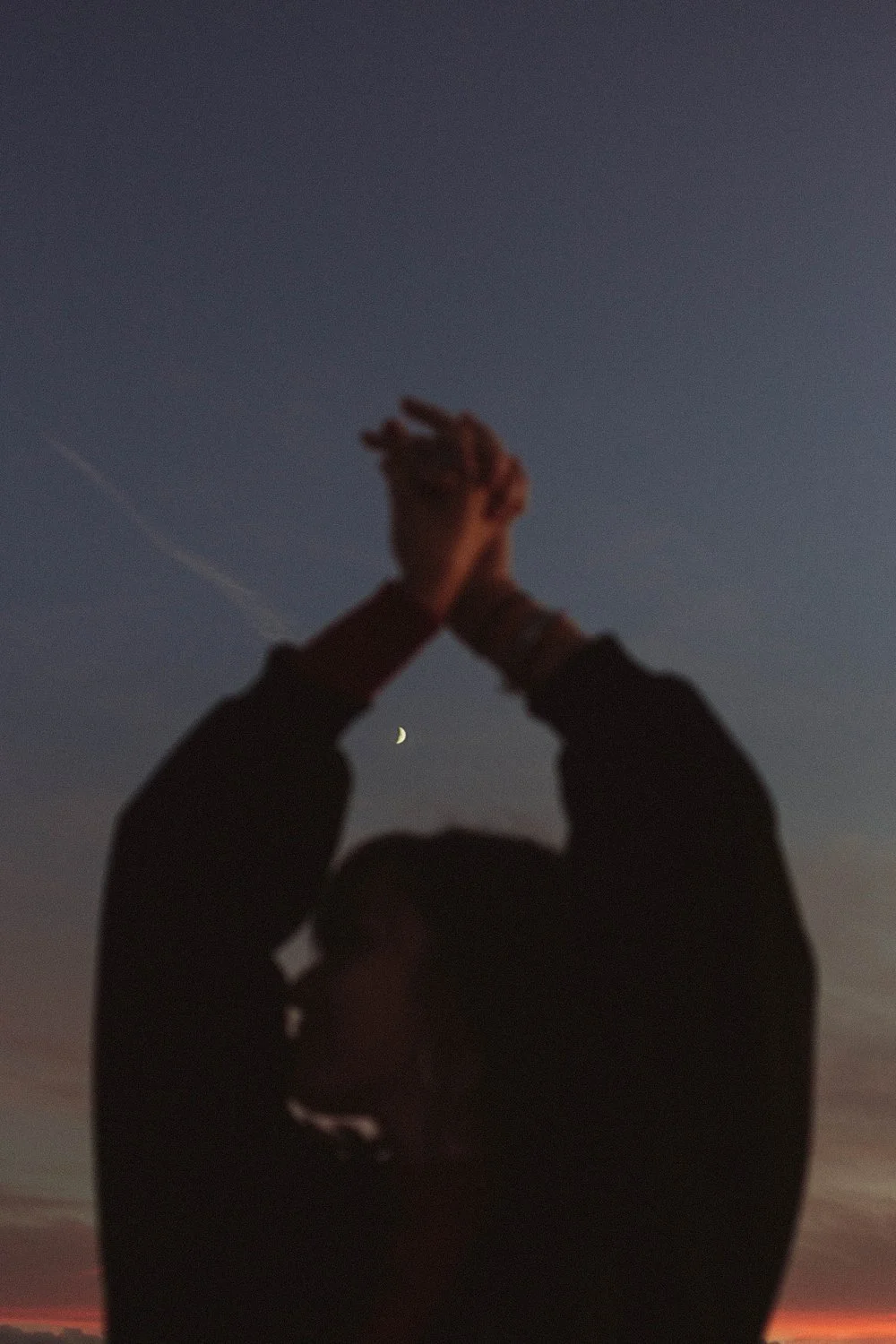 Silhouette of a woman at dusk with arms raised, a tiny crescent moon framed between her hands