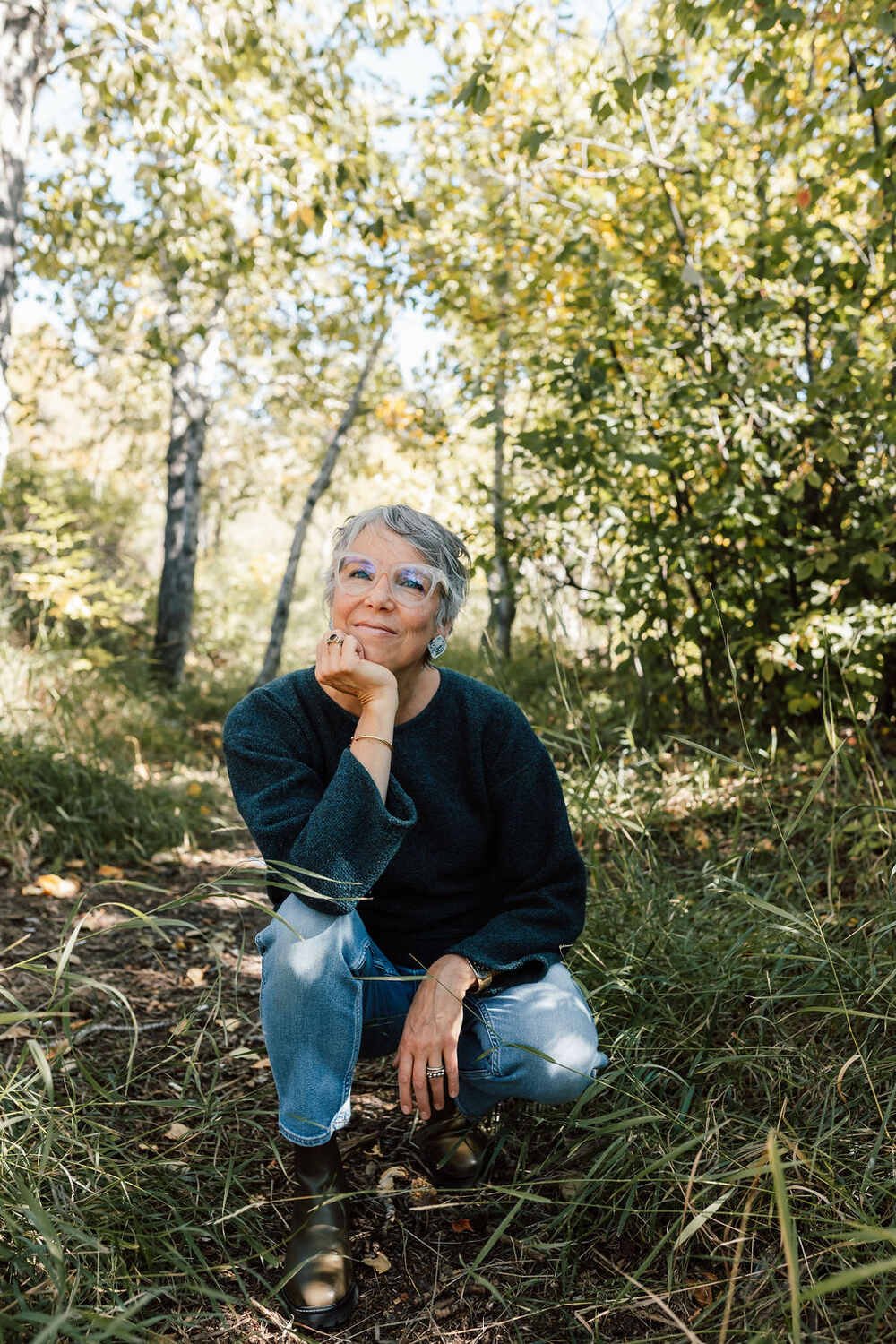 A woman with short gray hair, glasses, and jewelry squats on a forest trail, surrounded by trees with yellow and green leaves, posing with her chin resting on her hand and smiling.