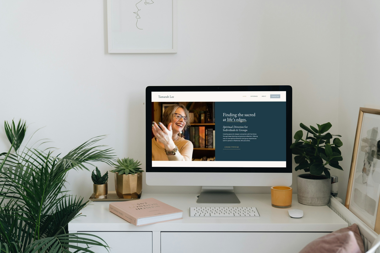 A clean white desk with potted plants, a closed book, a computer monitor displaying a website of Tamarah Lee with a woman smiling, and a framed artwork on a small table to the right.