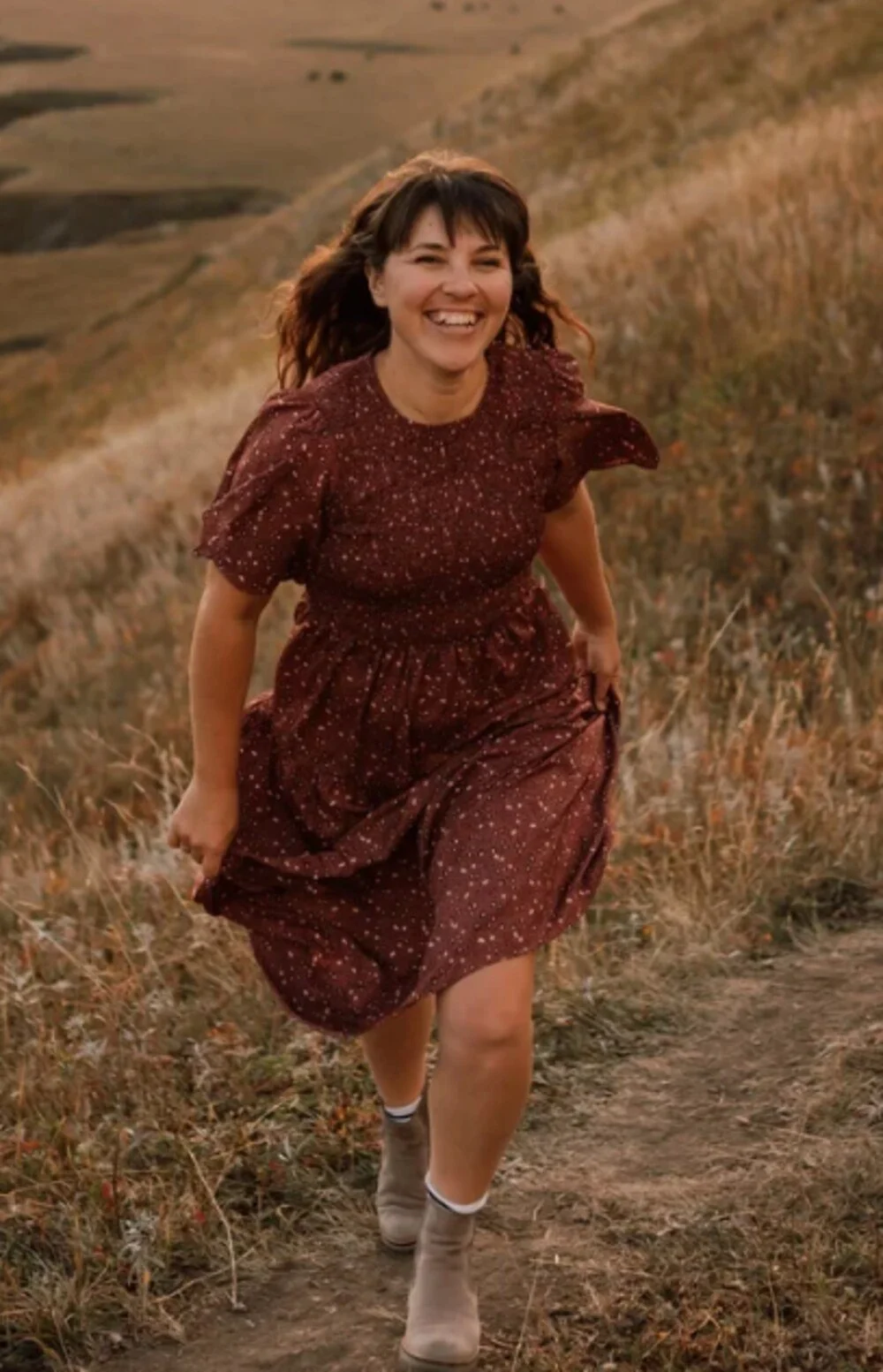 A woman smiling and running along a grassy trail in an open field with rolling hills in the background.