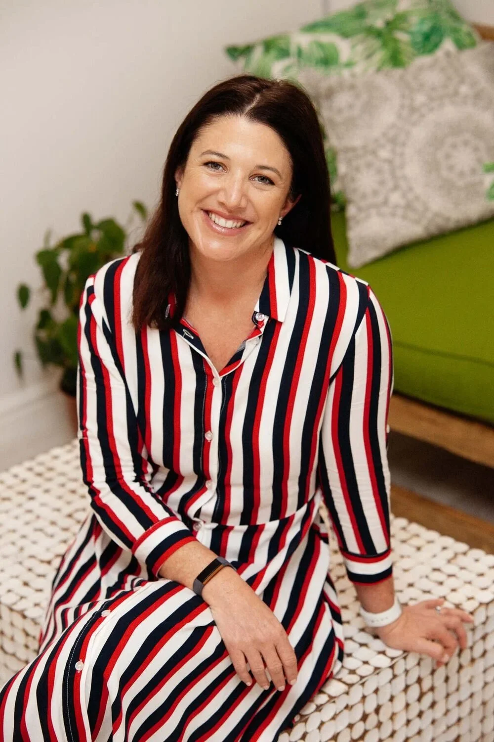 A woman with dark brown hair and a bright smile, wearing a red, white, and blue striped dress, sitting on a textured white surface in a room with green accents, including a green cushion and a plant in the background.