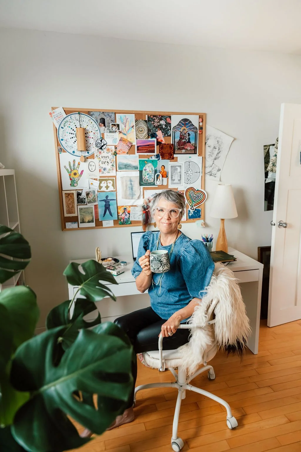 A woman with gray hair and glasses, wearing a denim top, smiling and holding a mug, sitting in a white office chair with a furry covering in a home office. Behind her, there is a bulletin board filled with colorful art, notes, and decorations, a white desk with supplies, and a lamp. A large green plant partially obstructs the view in the foreground.