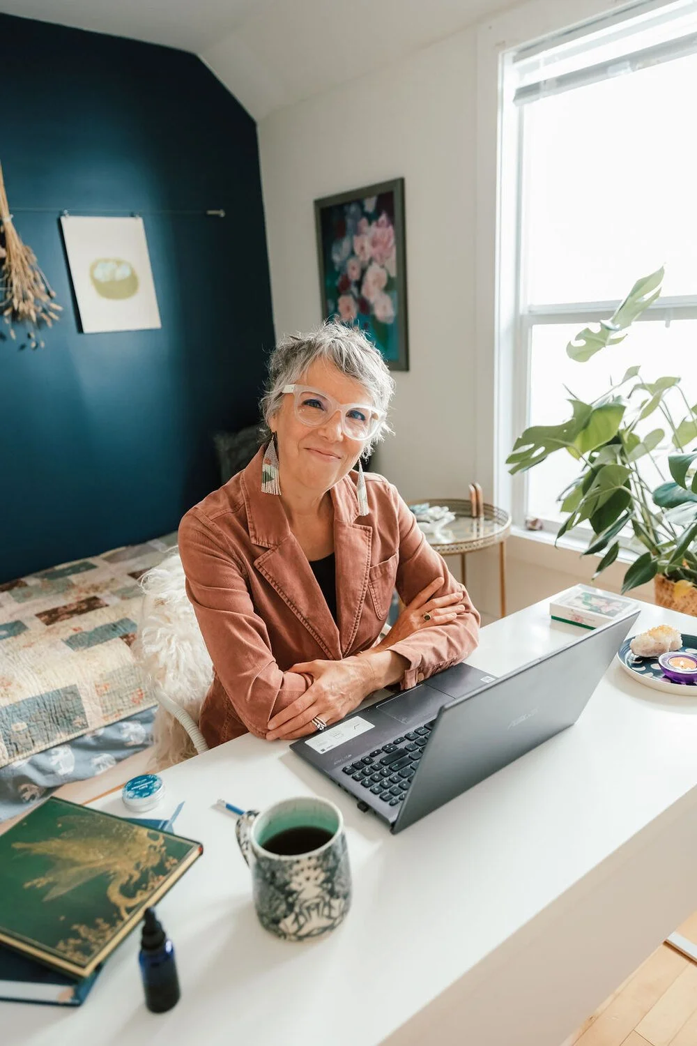 A woman with short gray hair, wearing glasses and earrings, sitting at a white desk with a laptop, a cup of coffee, and various items, in a bright room with a window and a green plant.