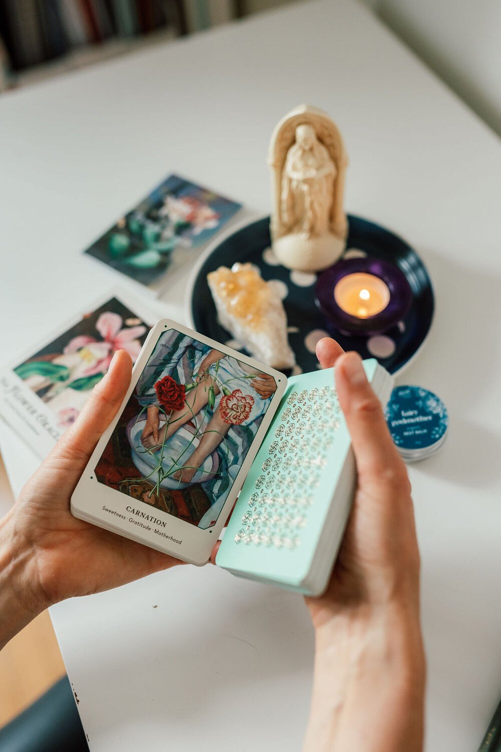 A person holding a deck of tarot cards, with a tarot card titled 'CARNATION: Sweetness, Gratitude, Motherhood' visible, and other cards and objects like a candle, religious figurine, and photographs on a table in the background.