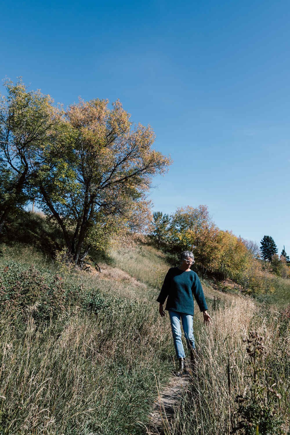 A woman with gray hair wearing glasses, a dark blue sweater, light blue jeans, and black boots walking through a grassy trail in a hilly area with trees showing fall foliage under a clear blue sky.