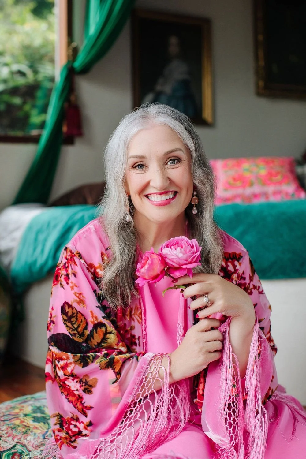 Portrait of Julie Parker with silver hair in a vibrant pink floral robe, holding pink roses warmly offering her praise of Moon Pause
