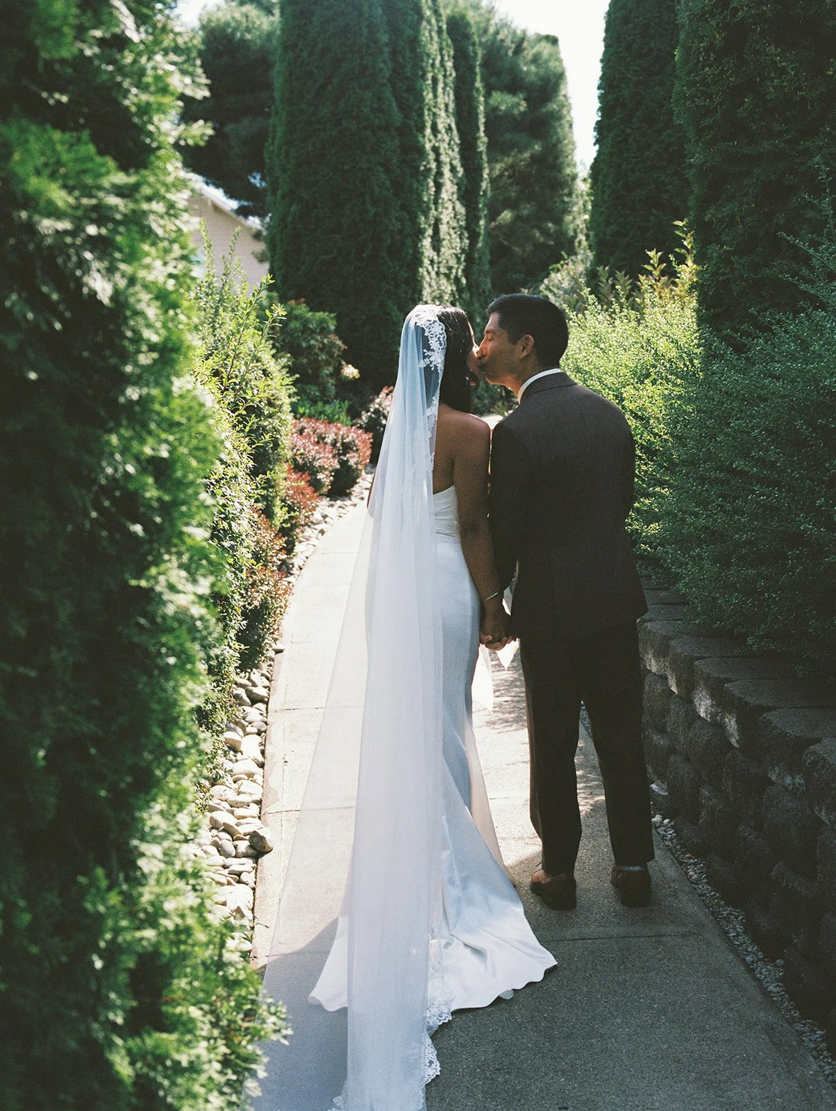 Bride and Groom walking away from the camera. They pause to kiss as they are surrounded by greenery hedges and soft lighting.