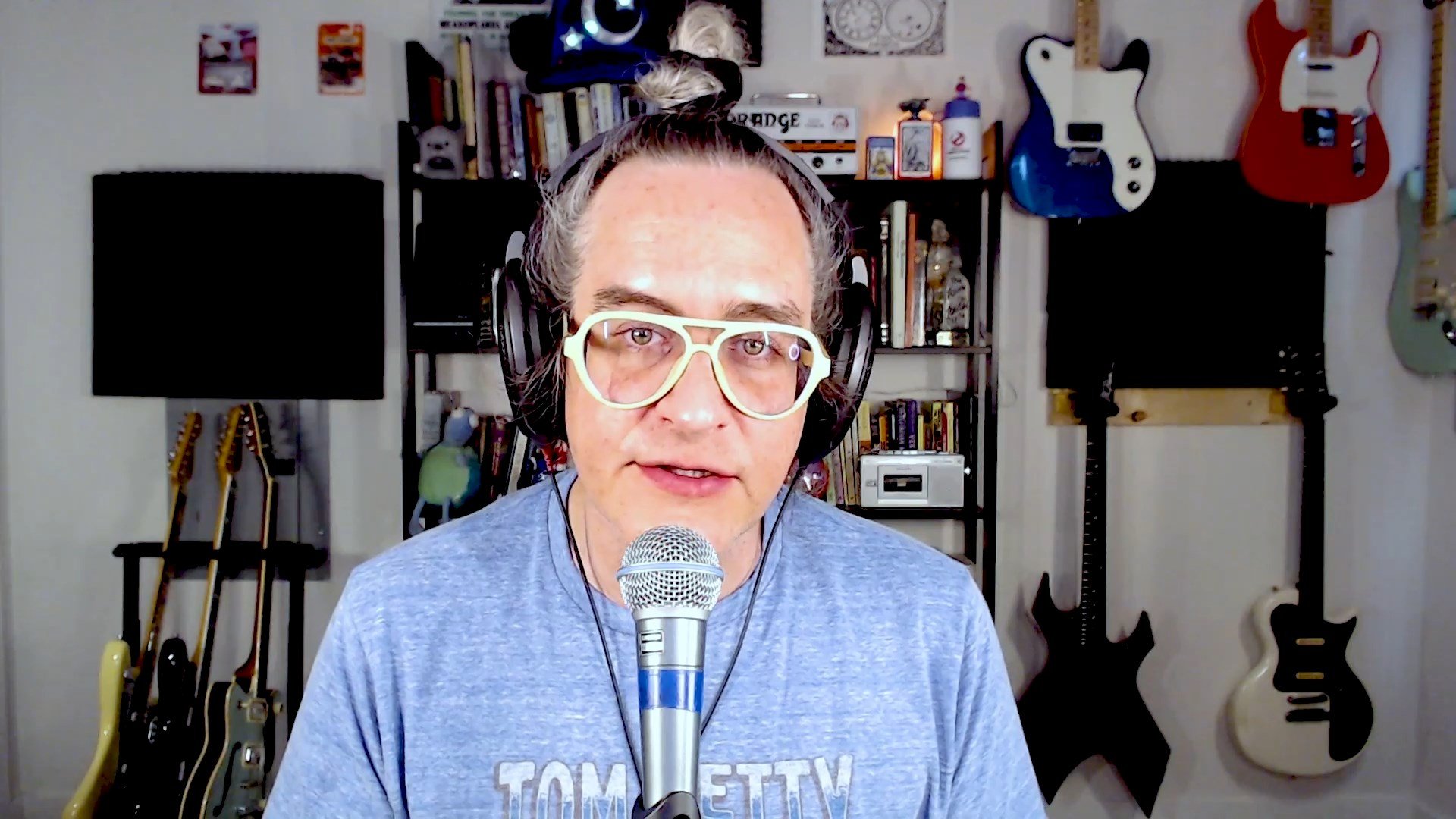 A crystal clear image of a man sitting in front of a wonderful backdrop of books, little trinkets and knick-knacks and electric guitars. You can also see an Orange Tiny Terror Amplifier, and a Disneyland Sorcerer Mickey hat with its stars aglow