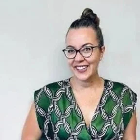A smiling woman with glasses and dark hair tied up, wearing a green patterned top.
