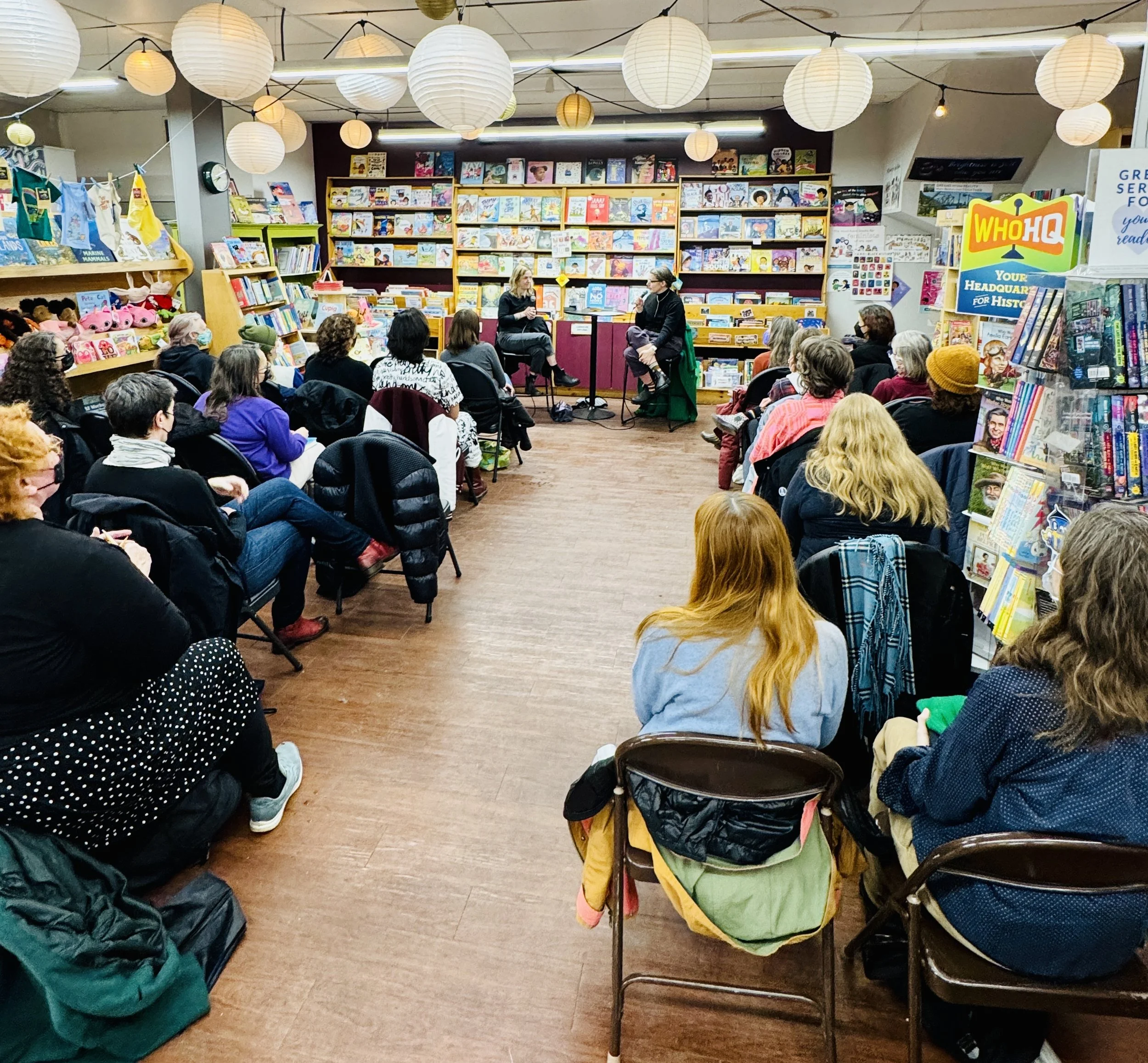 A book reading event in a bookstore with an audience listening to two women speaking in front of shelves filled with children's books, surrounded by paper lanterns hanging from the ceiling.