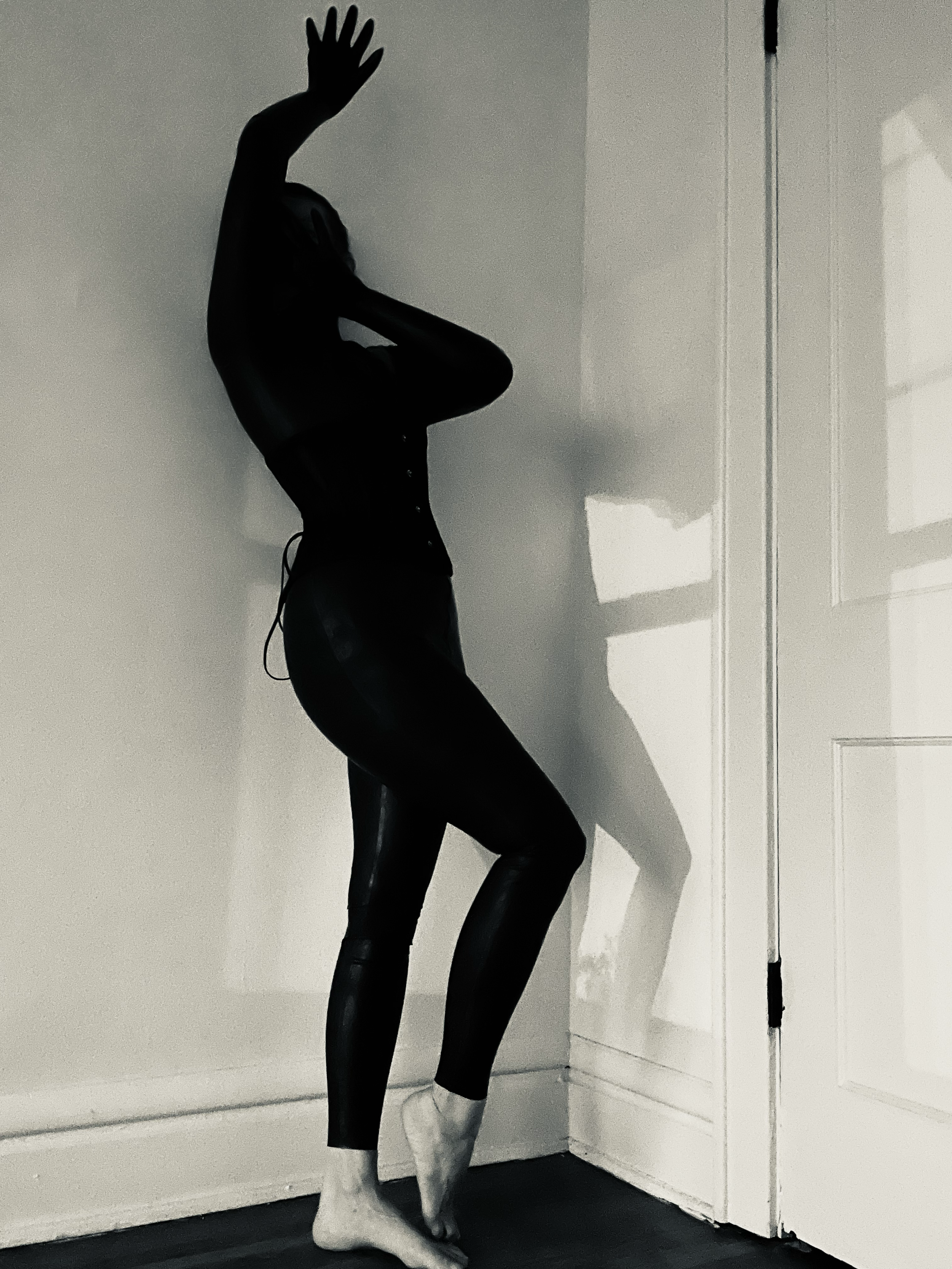 A black mannequin posed with one hand raised and the other touching the neck, standing barefoot indoors near a white wall and door, with shadows cast on the wall.