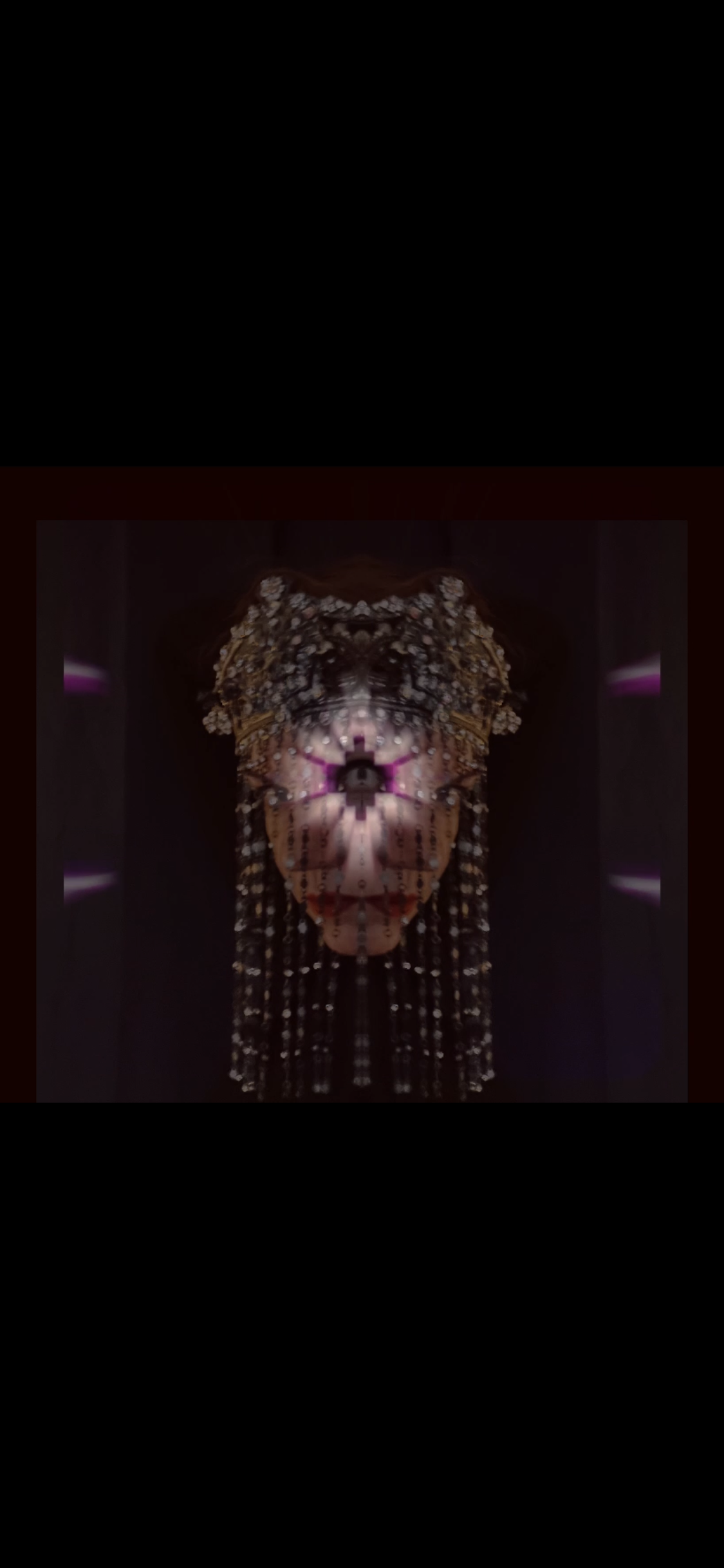 A person dressed in an elaborate costume with a headpiece made of flowers and beads, standing in front of a dark background with purple lights.