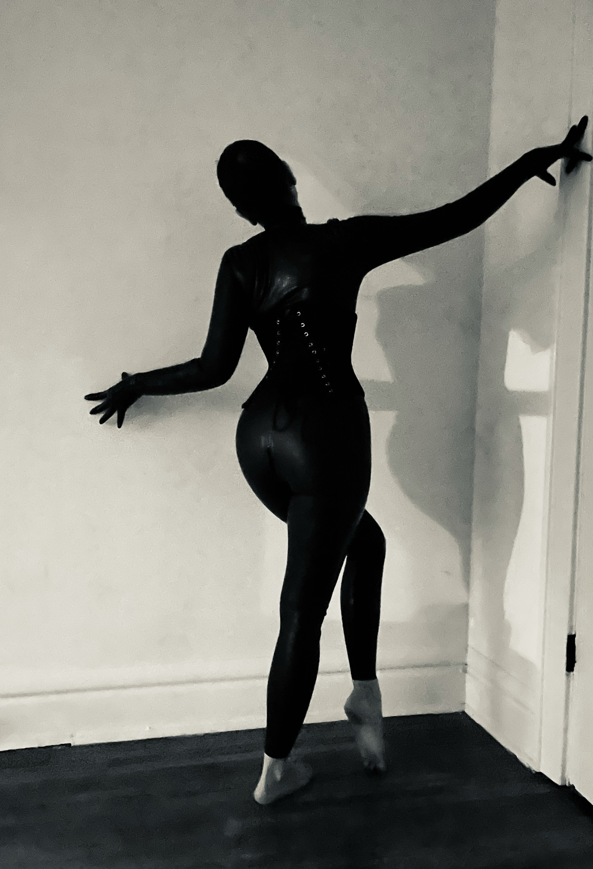 Silhouette of a woman in black leather clothing and high heels, standing against a plain wall, with one arm extended.