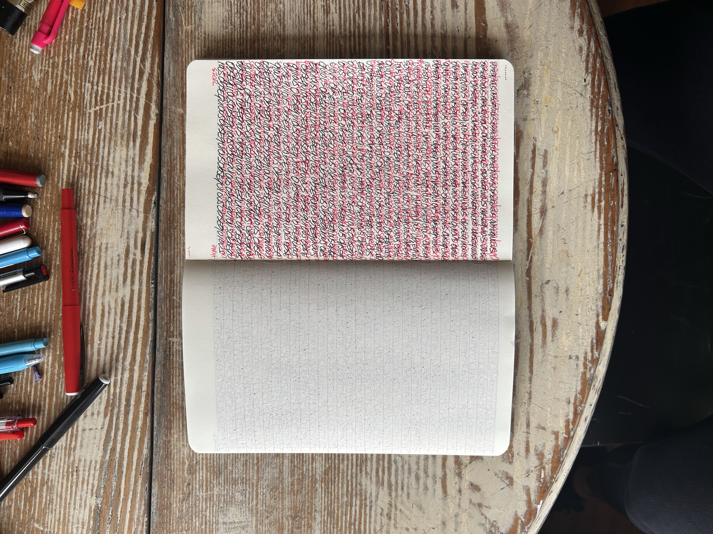 Open notebook with densely written text on the top page and blank grid-lined page below on a wooden table surrounded by colored pens and markers.