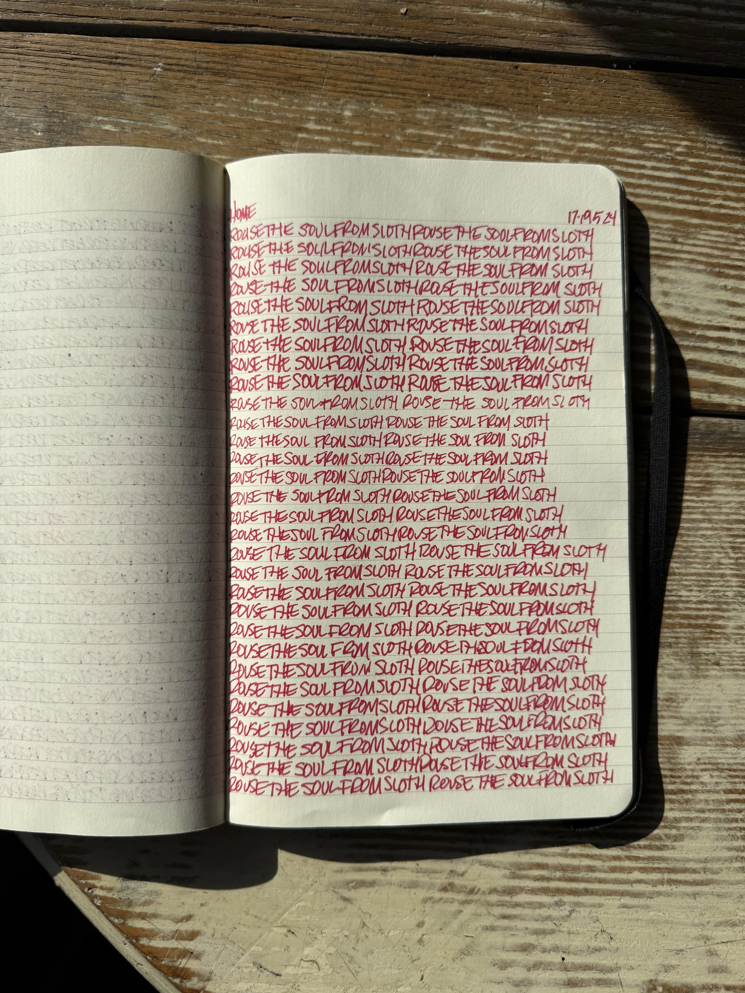 A spiral notebook open on a wooden surface. The left page is blank, while the right page contains handwritten text in red ink. The writing is repeated lines of the phrase: 'RAISE THE SOUL FROM SLOTH' and 'RAISE THE SOUL FROM SLOTH FROM SLOTH' among o