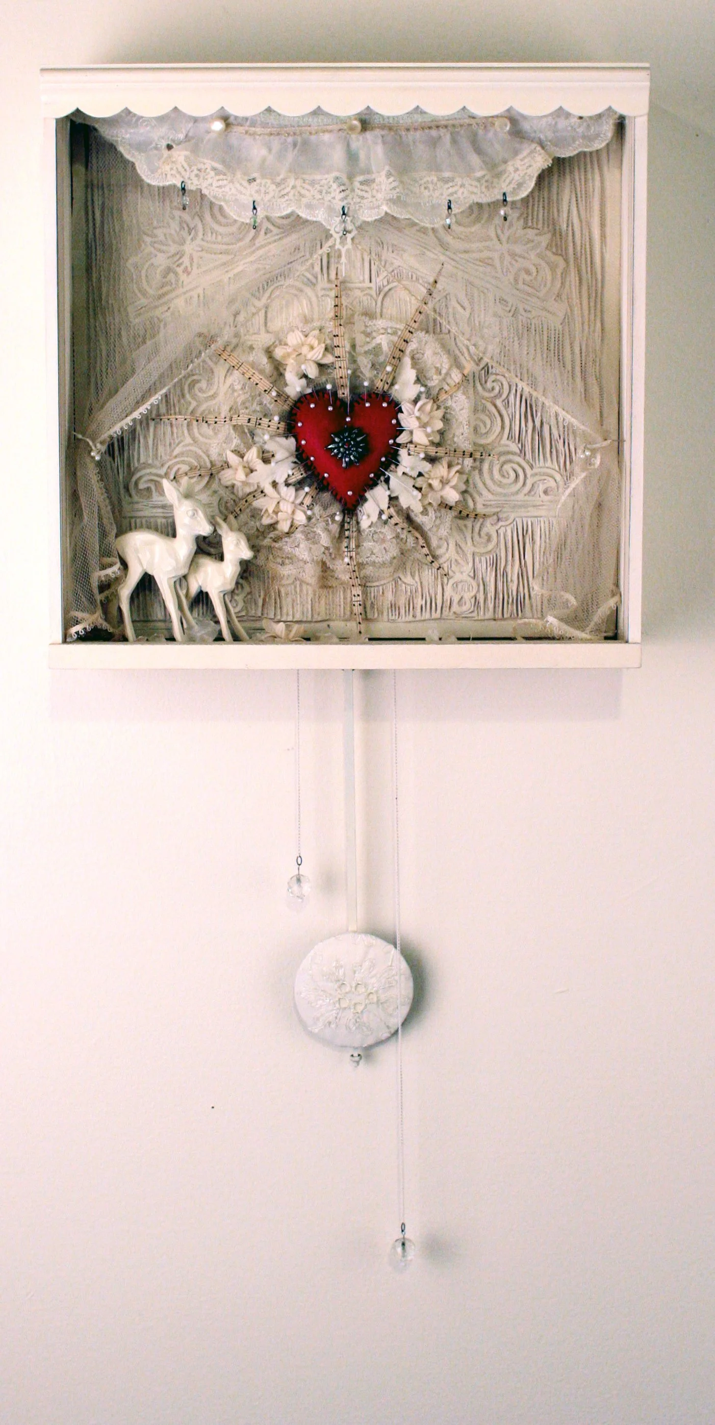 Decorative shadow box with lace and lace cardigan, white jewelry, surrounded by white ceramic deer, featuring a heart with radiating sticks and flowers, with hanging crystal and circular ornament.
