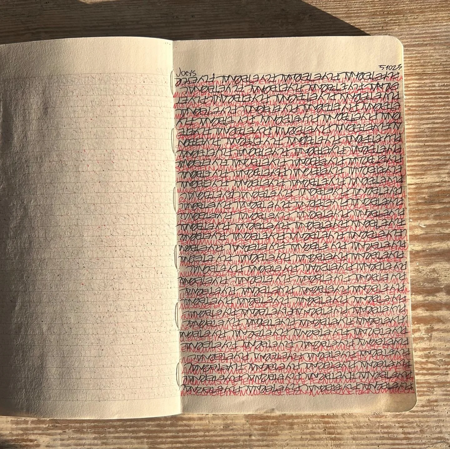 An open notebook on a wooden surface with scribbles and overlapping writings in black and red ink, filling the right page, while the left page is blank.
