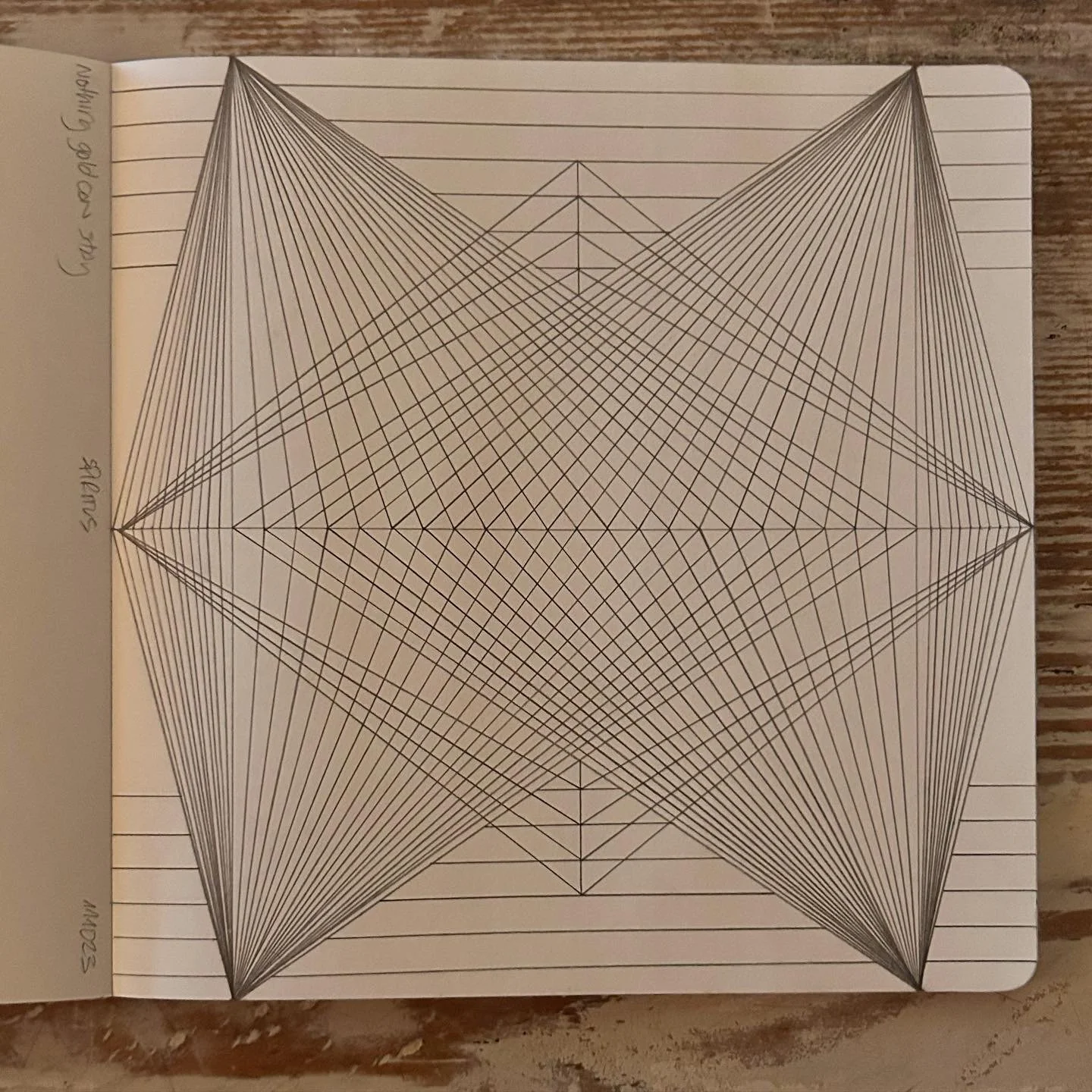 A geometric drawing with intersecting straight lines forming a complex, symmetrical starburst pattern on lined paper placed on a wooden surface.