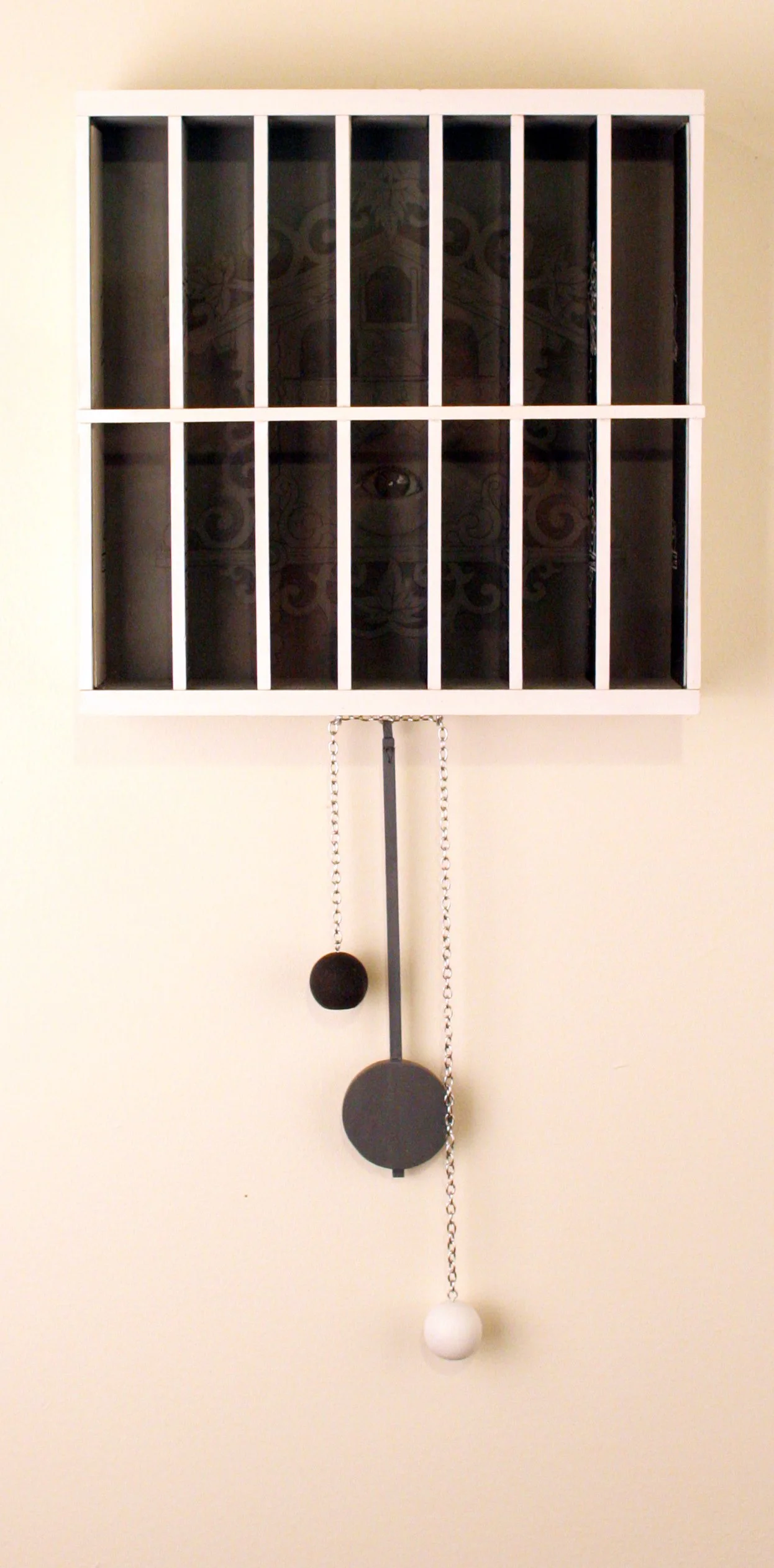 A large wall clock designed like a jail cell with white bars, black background, and a dark face. The clock has three chains with colored balls hanging from it.