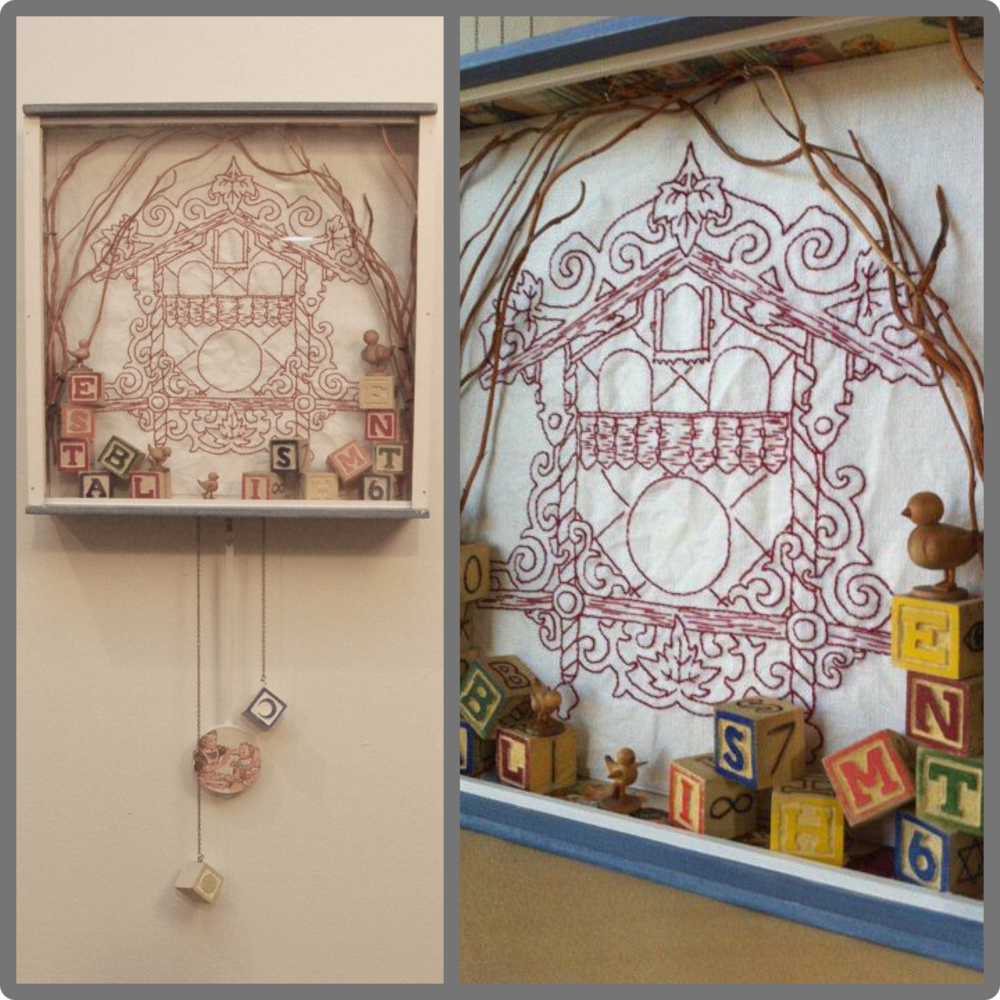Decorative shadow box featuring a house outline drawn on fabric, surrounded by vine-like shapes, with wooden duck figurines and colorful alphabet blocks arranged at the bottom.