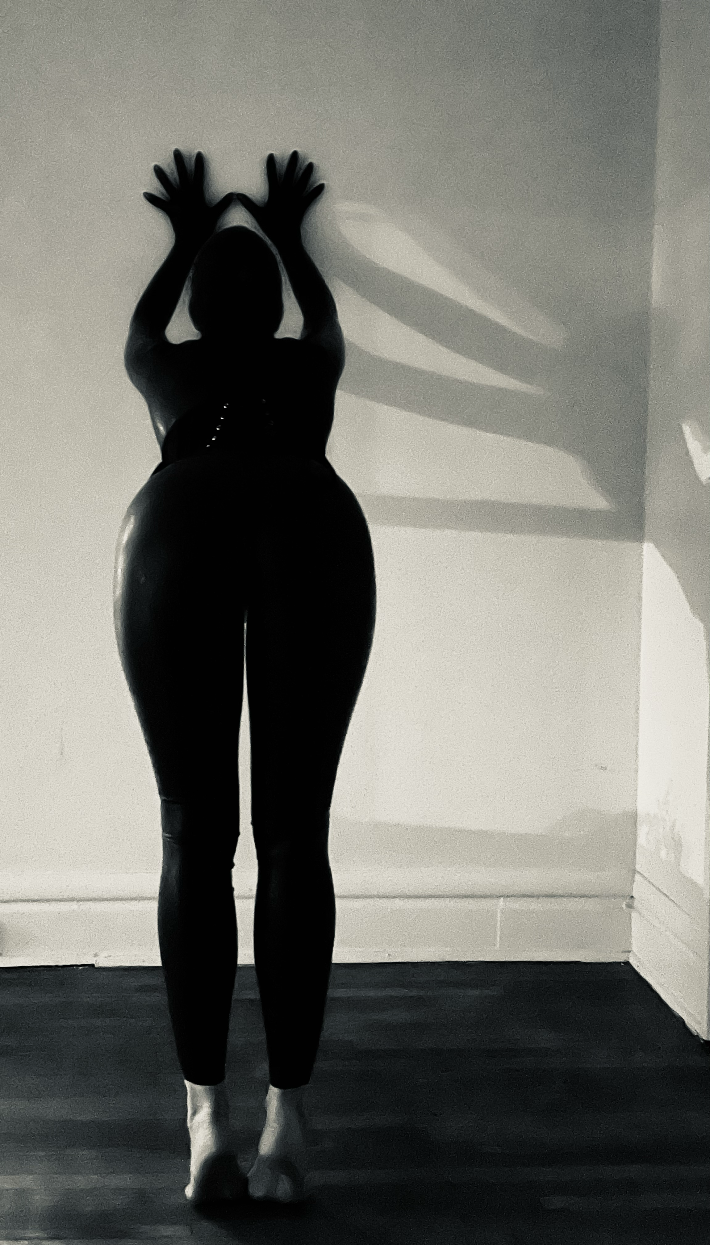 Silhouette of a person standing on a floor, facing a wall, with their hands raised above their head and their shadow cast on the wall.