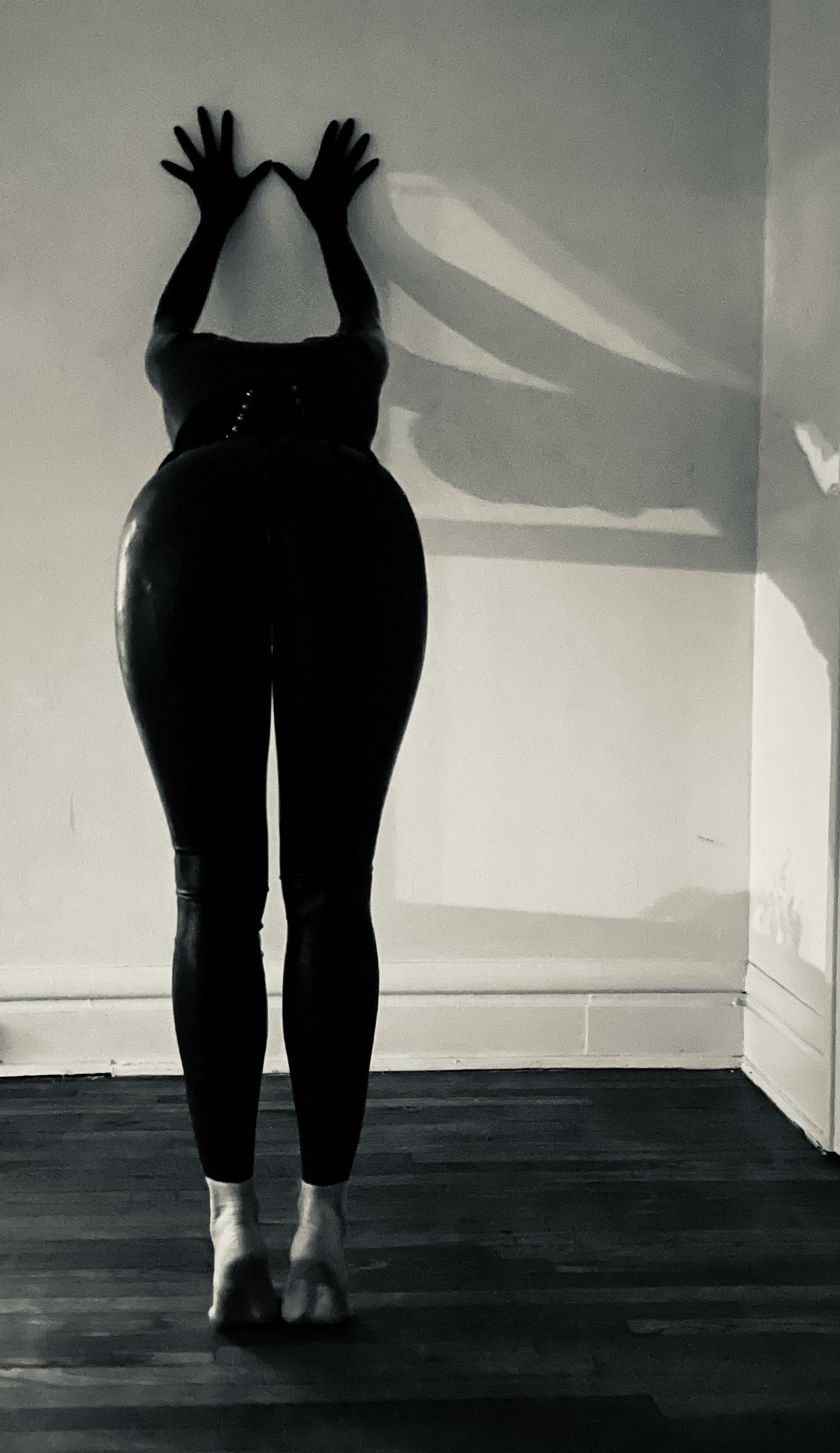 A person dressed in tight black clothing, facing a white wall, with hands pressed against the wall and shadow cast on the wall
