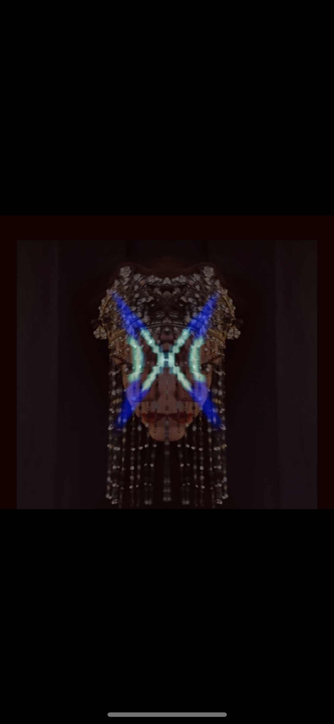 Blurred image of an illuminated display with a mirrored pattern resembling butterfly wings, featuring vibrant blue and green colors, likely at a nighttime event or exhibit.
