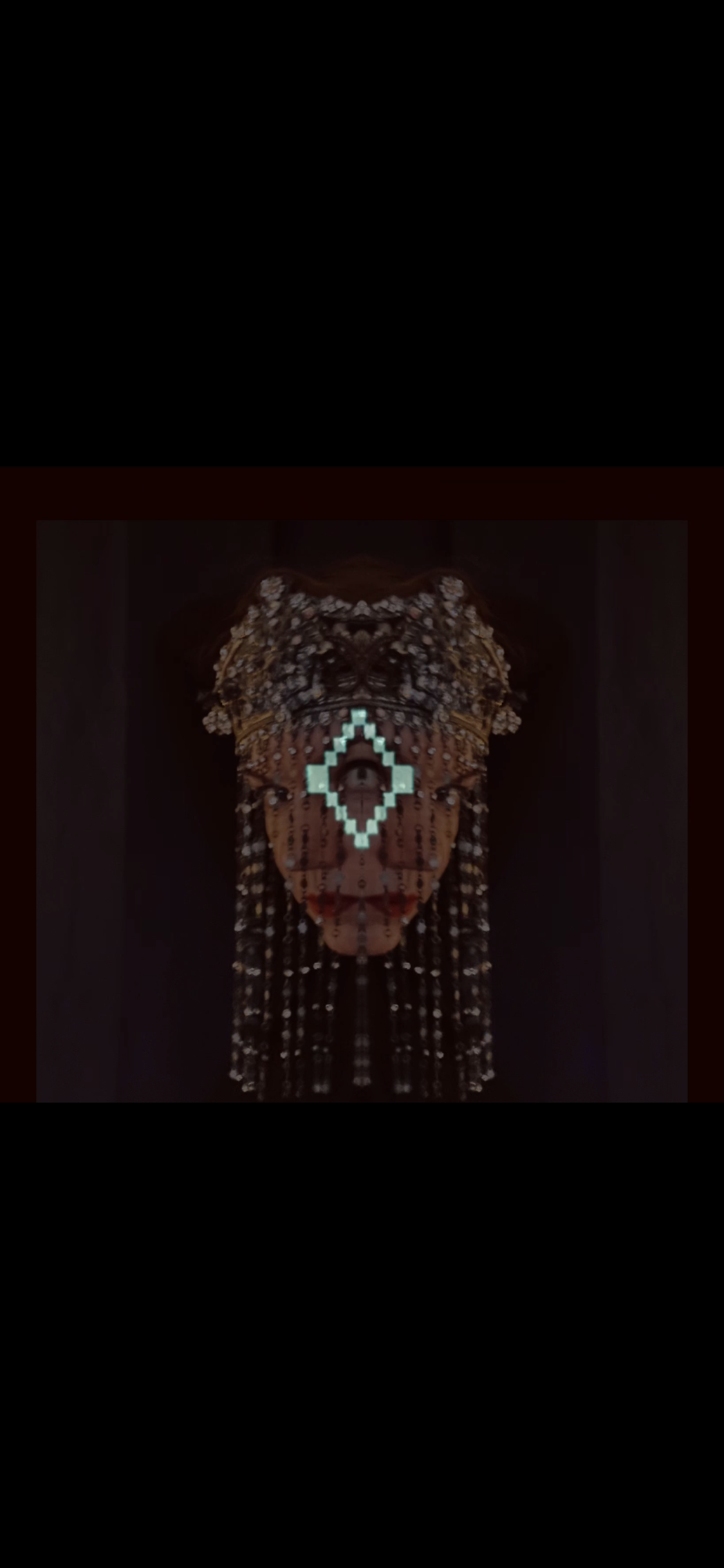 An artwork combining a digital and traditional mask with a human eye visible through a diamond-shaped digital frame, adorned with beads, shells, and feathers.