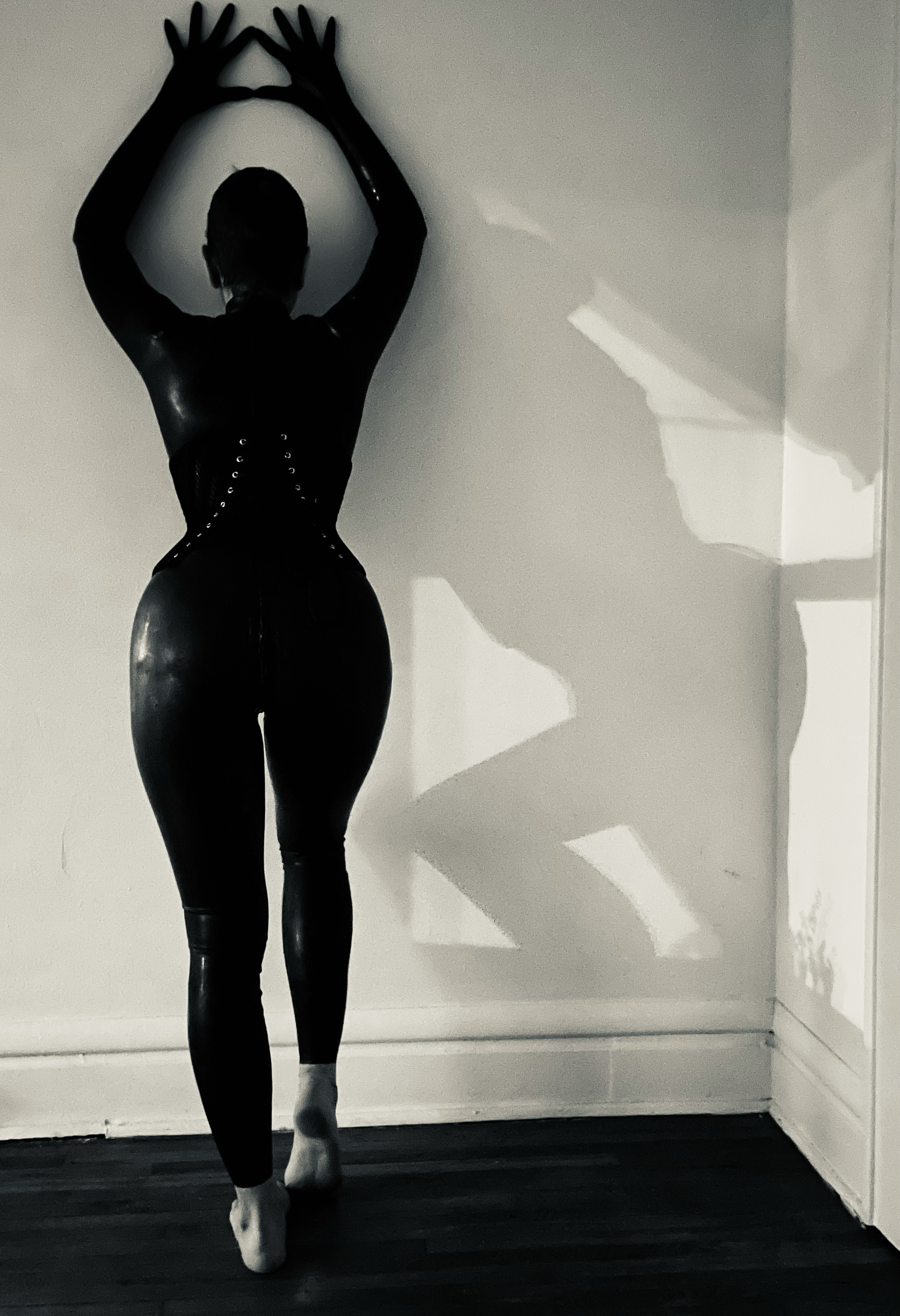 A person dressed in black latex clothing standing with their back to the camera, casting a shadow on the wall.