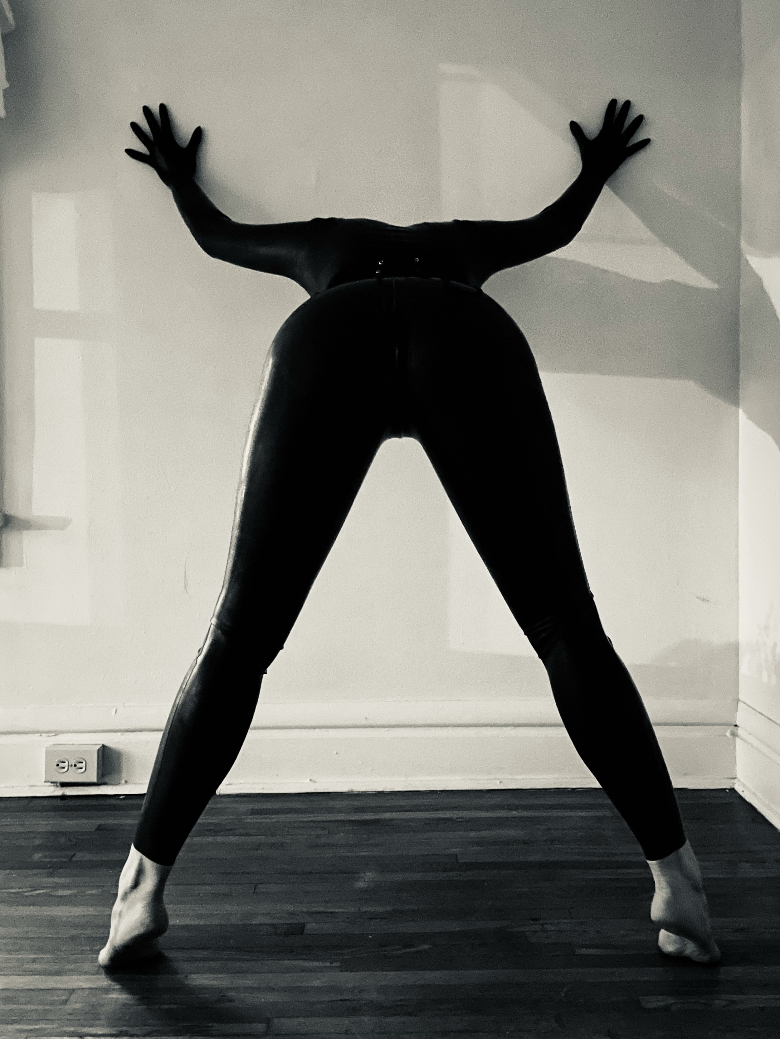 Silhouette of a person standing against a wall with arms extended upward, wearing tight clothing, with feet apart on a wooden floor.